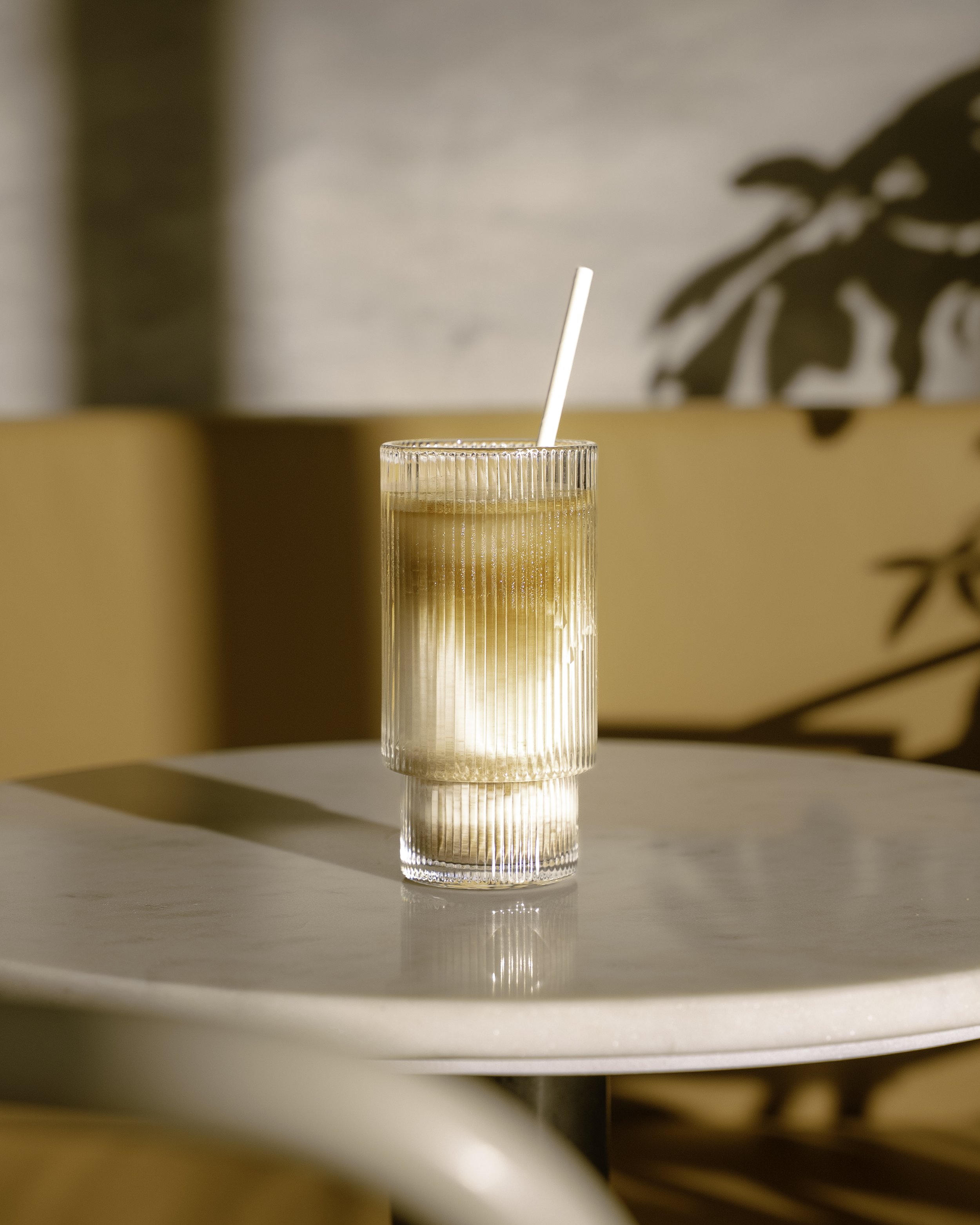 Un verre transparent avec une boisson glacée et une paille blanche, placé sur une table blanche dans un environnement lumineux et moderne.