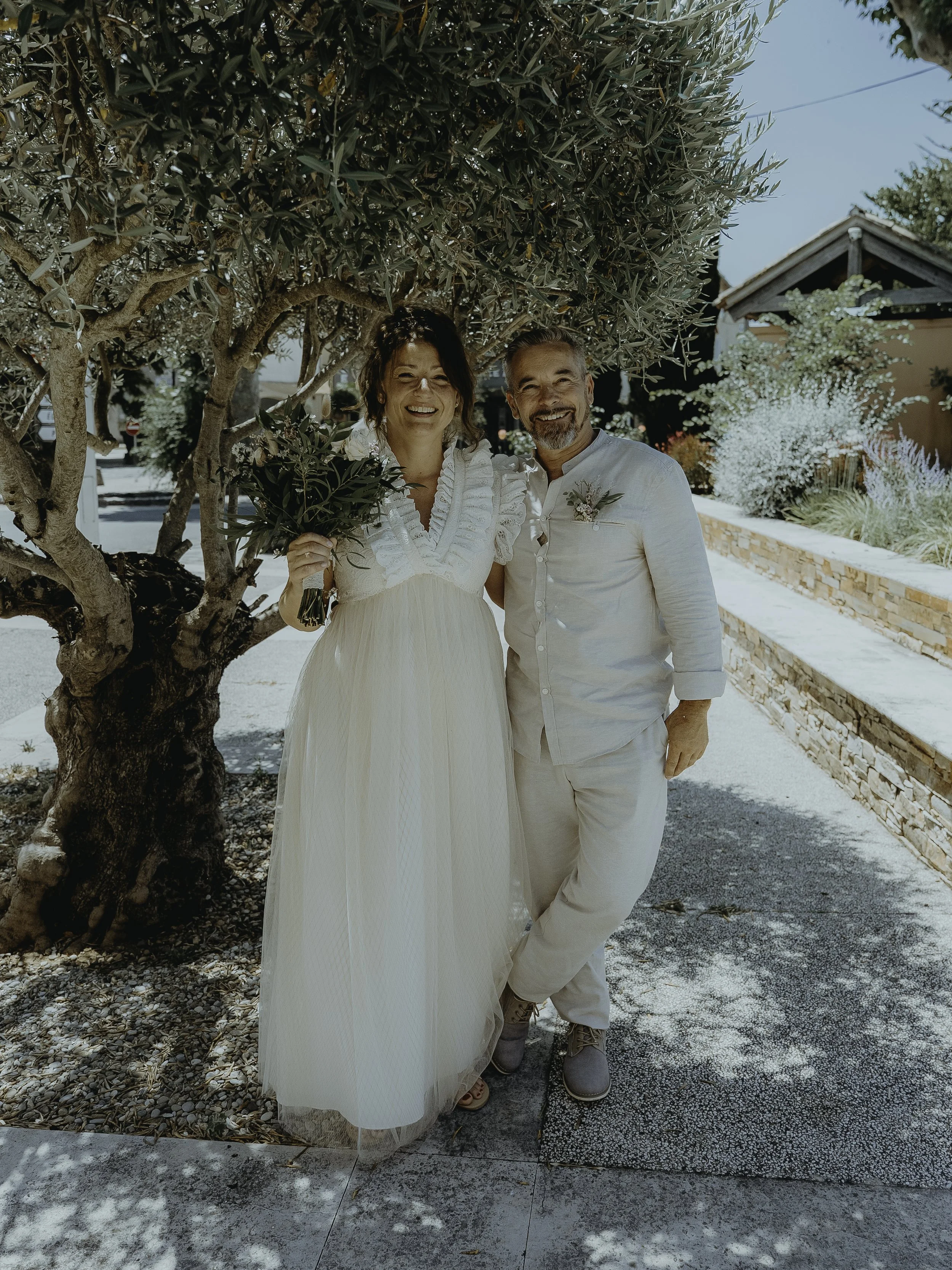 Un couple souriant en vêtements blancs, probablement lors d'un mariage, posant sous un arbre avec un cadre naturel et lumineux.