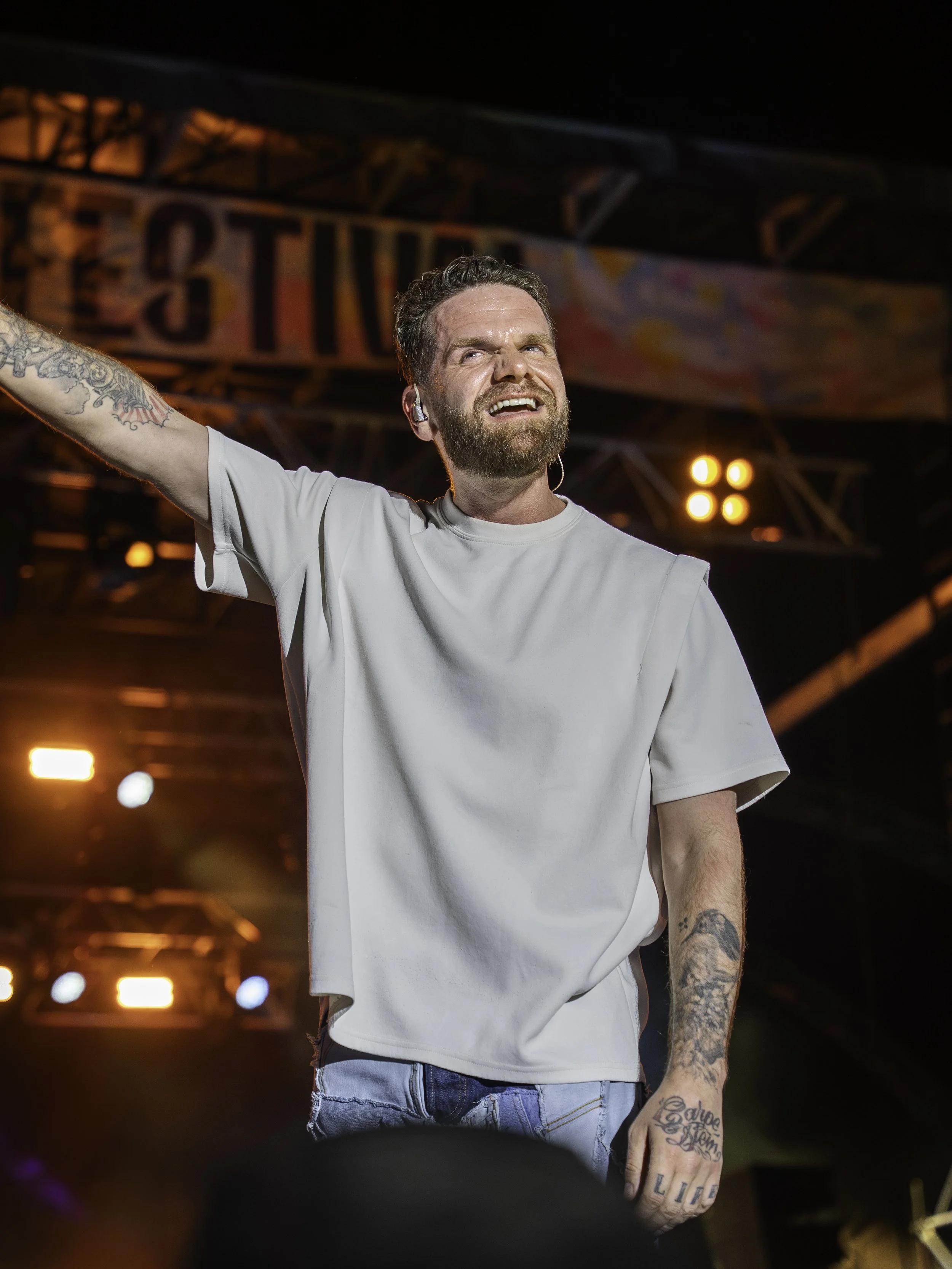 Un homme avec une barbe, en chemise blanche, tenant un micro et levant le bras, en train de performer sur scène lors d'un concert. La scène est éclairée avec des lumières de scène orange et bleues.