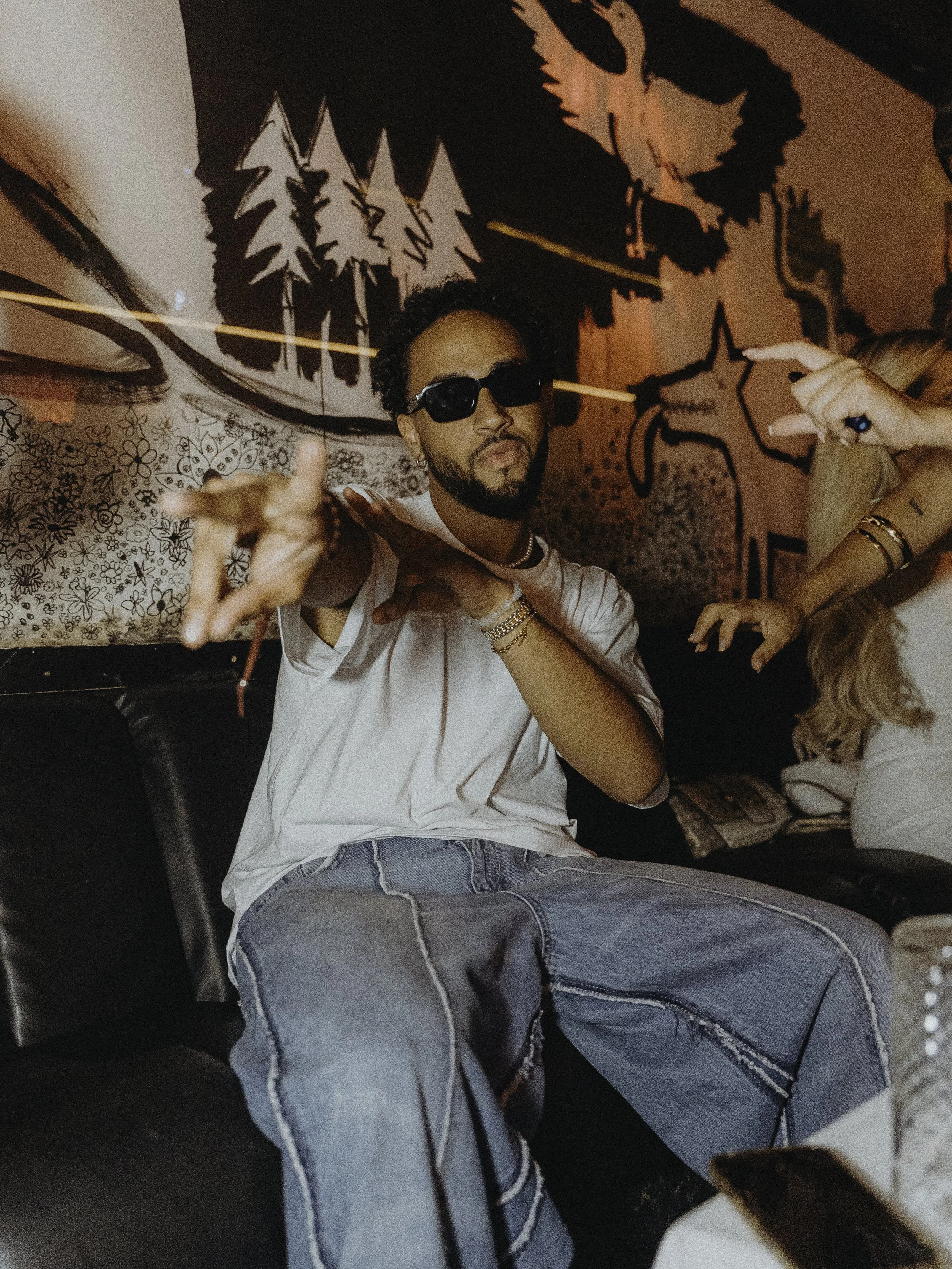 Un homme assis sur un canapé, portant des lunettes de soleil noires, un t-shirt blanc, des jeans déchirés, et plusieurs bracelets, fait un geste de la main vers l'objectif dans une pièce décorée avec un mur peint de dessins d'animaux et d'arbres.