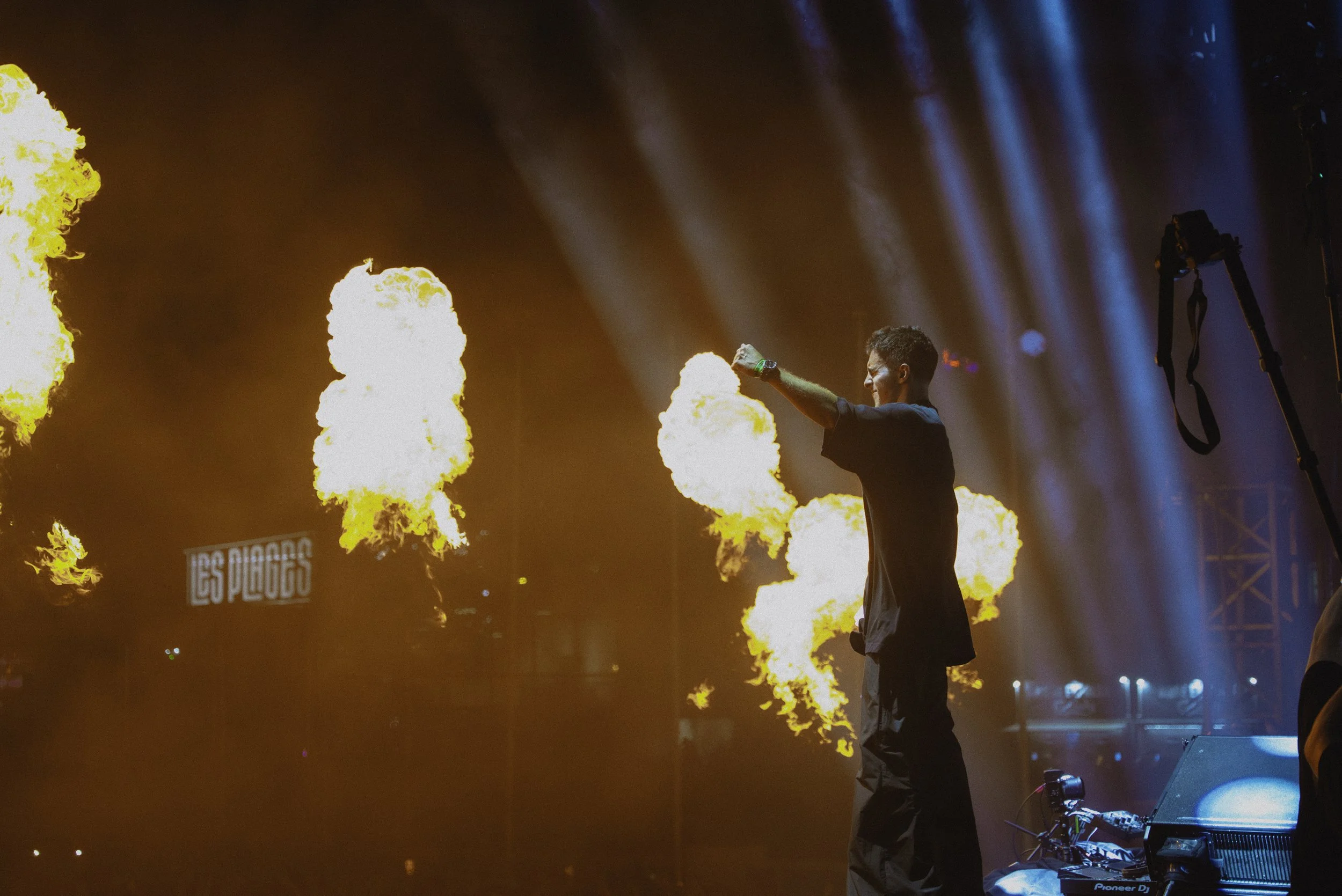 Un DJ sur scène avec des flammes en arrière-plan, illuminé par des projecteurs bleus, lors d'un concert de Les Pueblos.