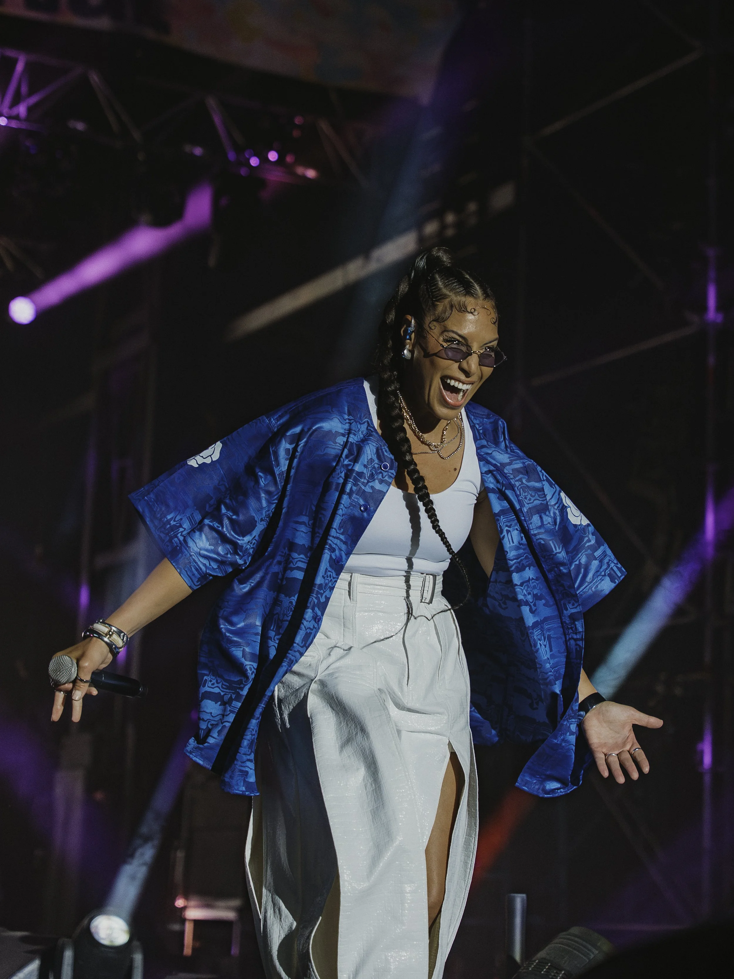 Une femme souriante chante en concert, portant une veste bleue, une jupe blanche fendue, des lunettes violettes, avec des lumières colorées en arrière-plan.