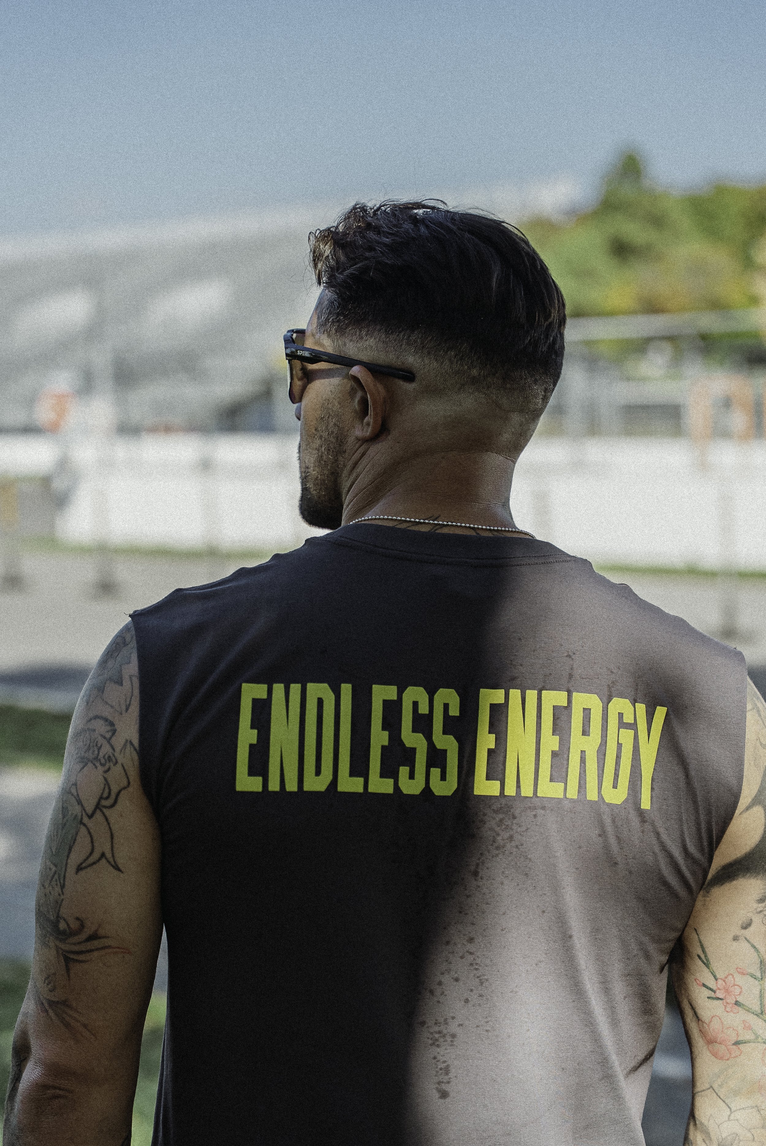 Un homme tatoué vu de dos, portant un t-shirt noir avec l'inscription « ENDLESS ENERGY » en lettres vertes, portant des lunettes de soleil, dans un environnement extérieur lors d'une journée ensoleillée.