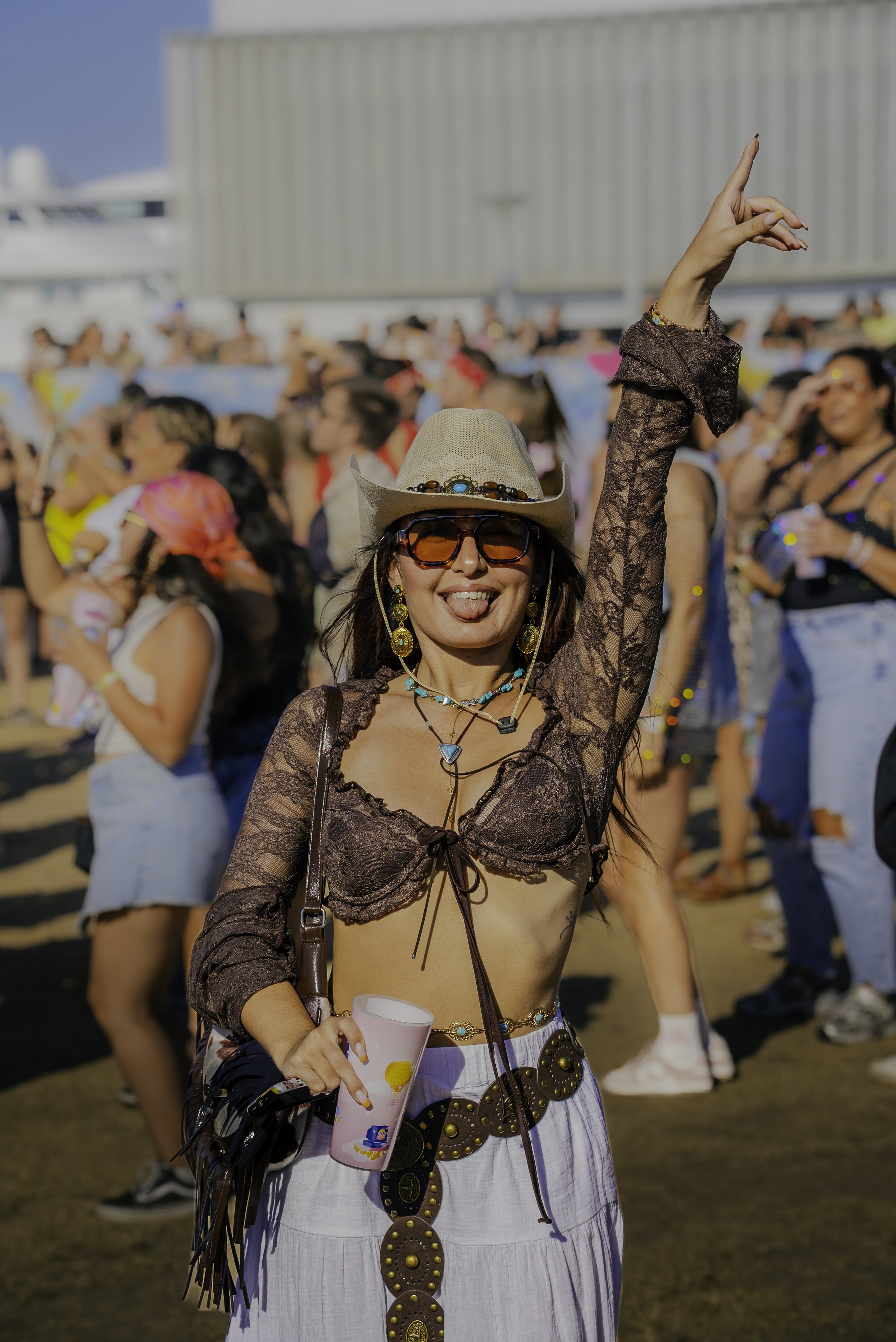 Une femme souriante portant une chemise en dentelle, un chapeau de cowboy, des lunettes noires, des boucles d'oreilles, un collier, un short blanc avec une ceinture large et un bracelet, tient une tasse dans sa main lors d'un festival en plein air av