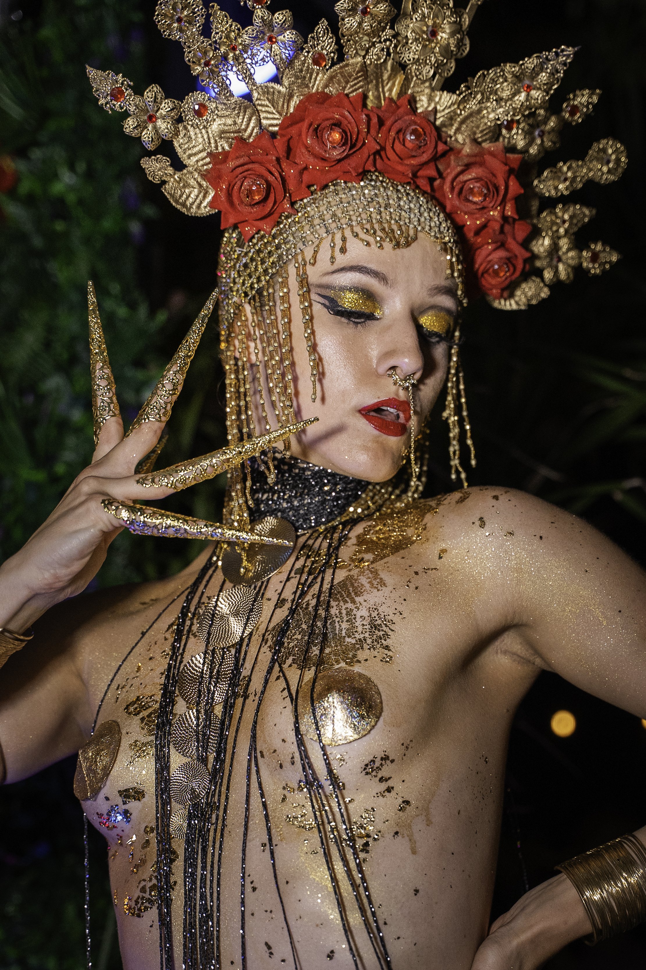 Une femme portant une coiffe ornementale décorée de roses rouges et de motifs dorés, avec un maquillage doré et rouge vif, des bijoux dorés, des décorations en or sur le corps, et des ongles ornés de bijoux.