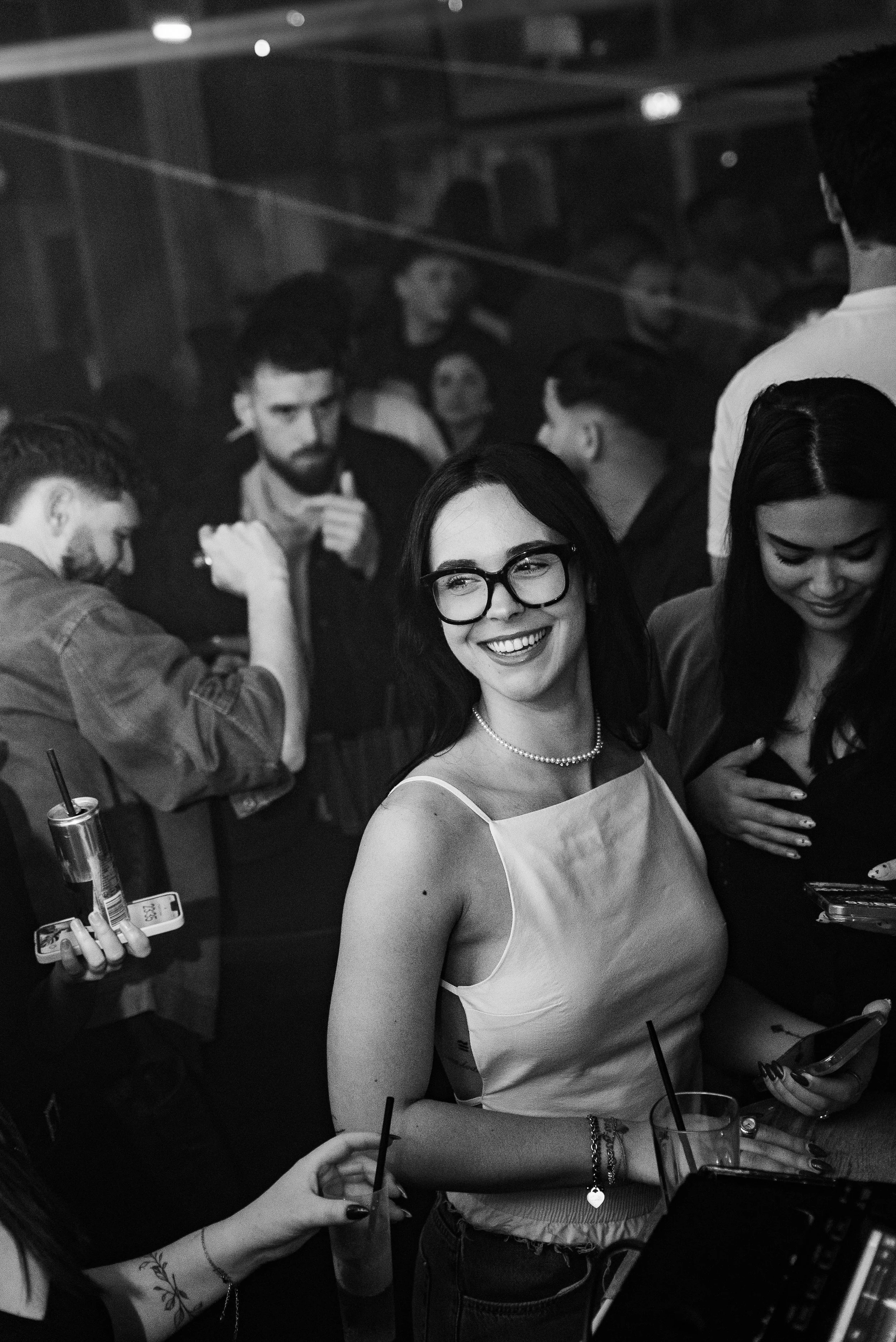 Fête en boîte de nuit avec jeunes adultes, une femme souriante portant des lunettes et un collier de perles, ambiance animée.