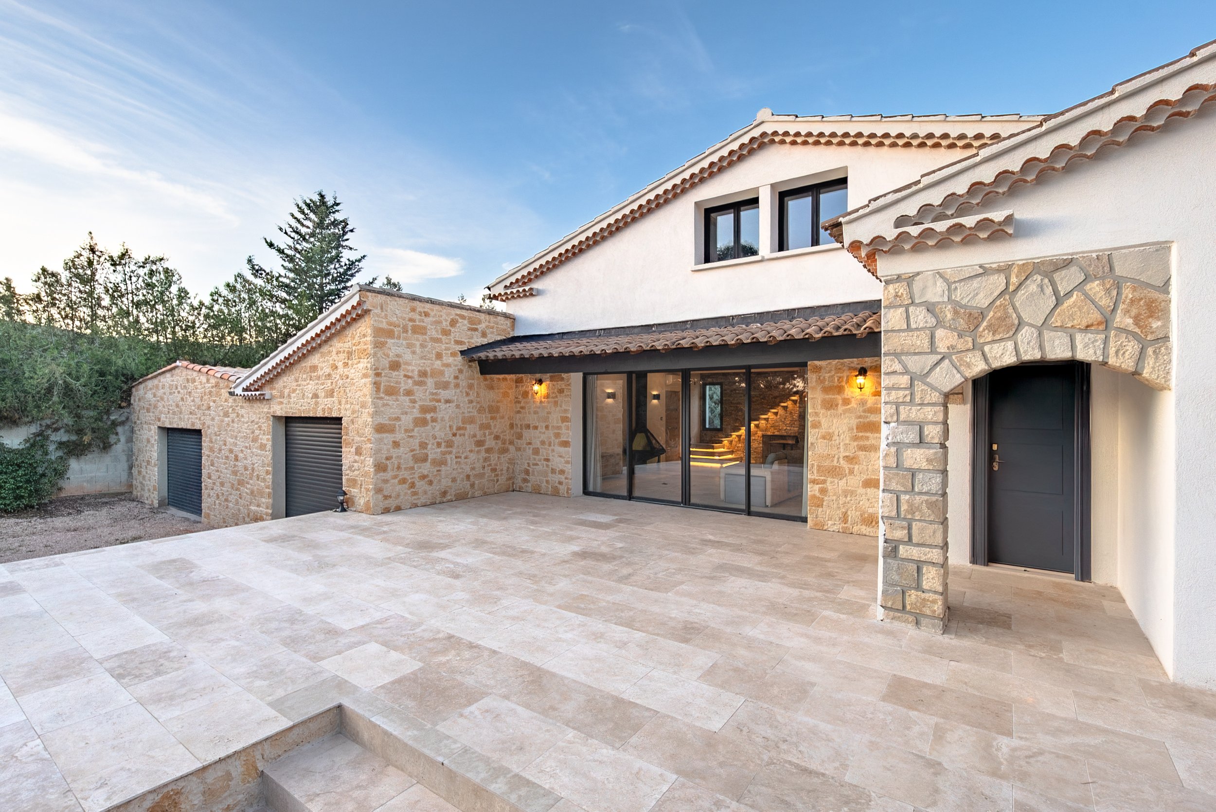 Vue extérieure d'une maison moderne avec murs en pierre et volets roulants noirs, grande terrasse en pierre et fenêtres en verre.