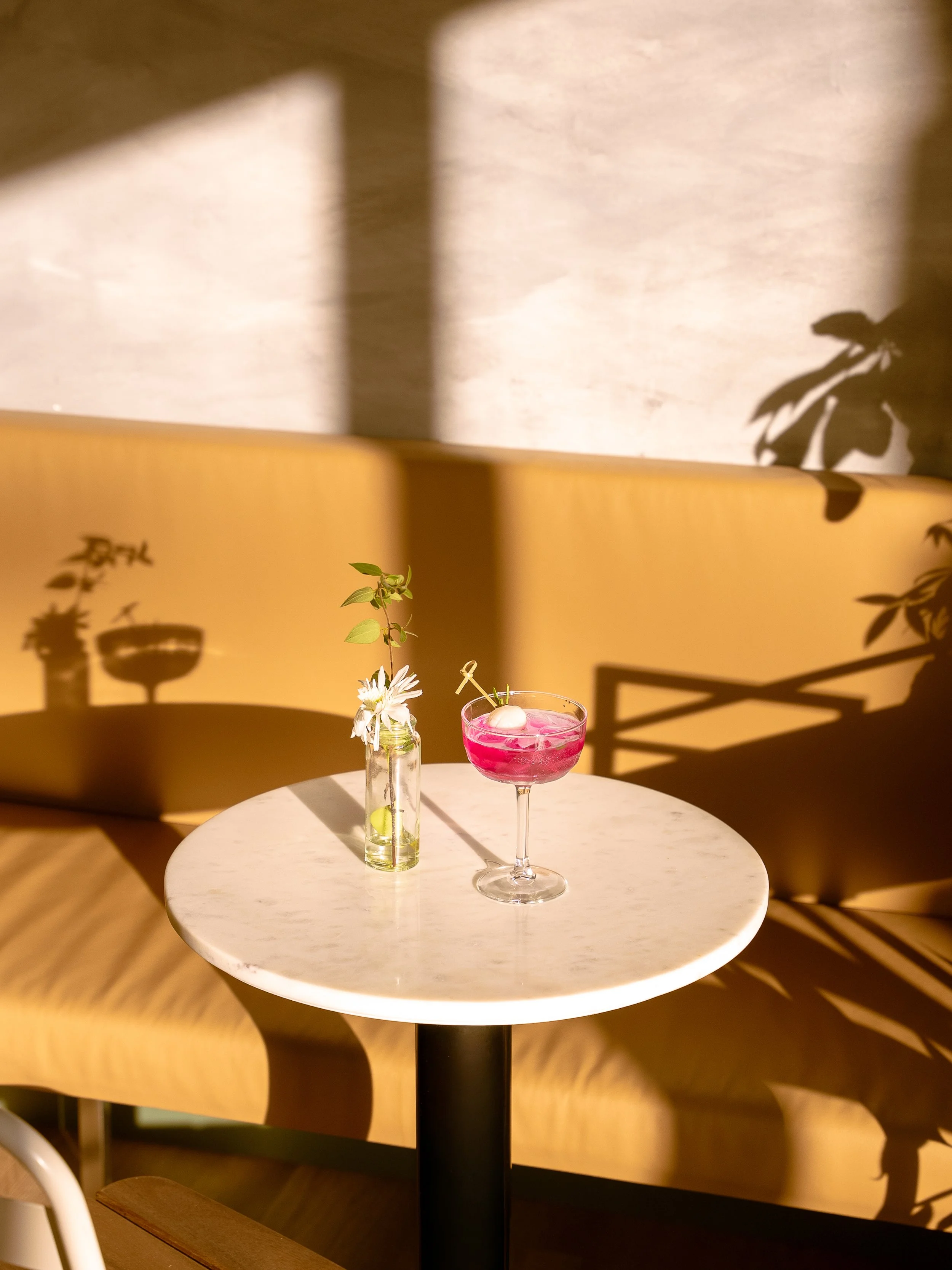 Un verre à cocktail rose sur une table ronde en marbre avec une petite bouteille en verre contenant une fleur et des feuilles. Shadows de plantes projetés sur un mur beige.