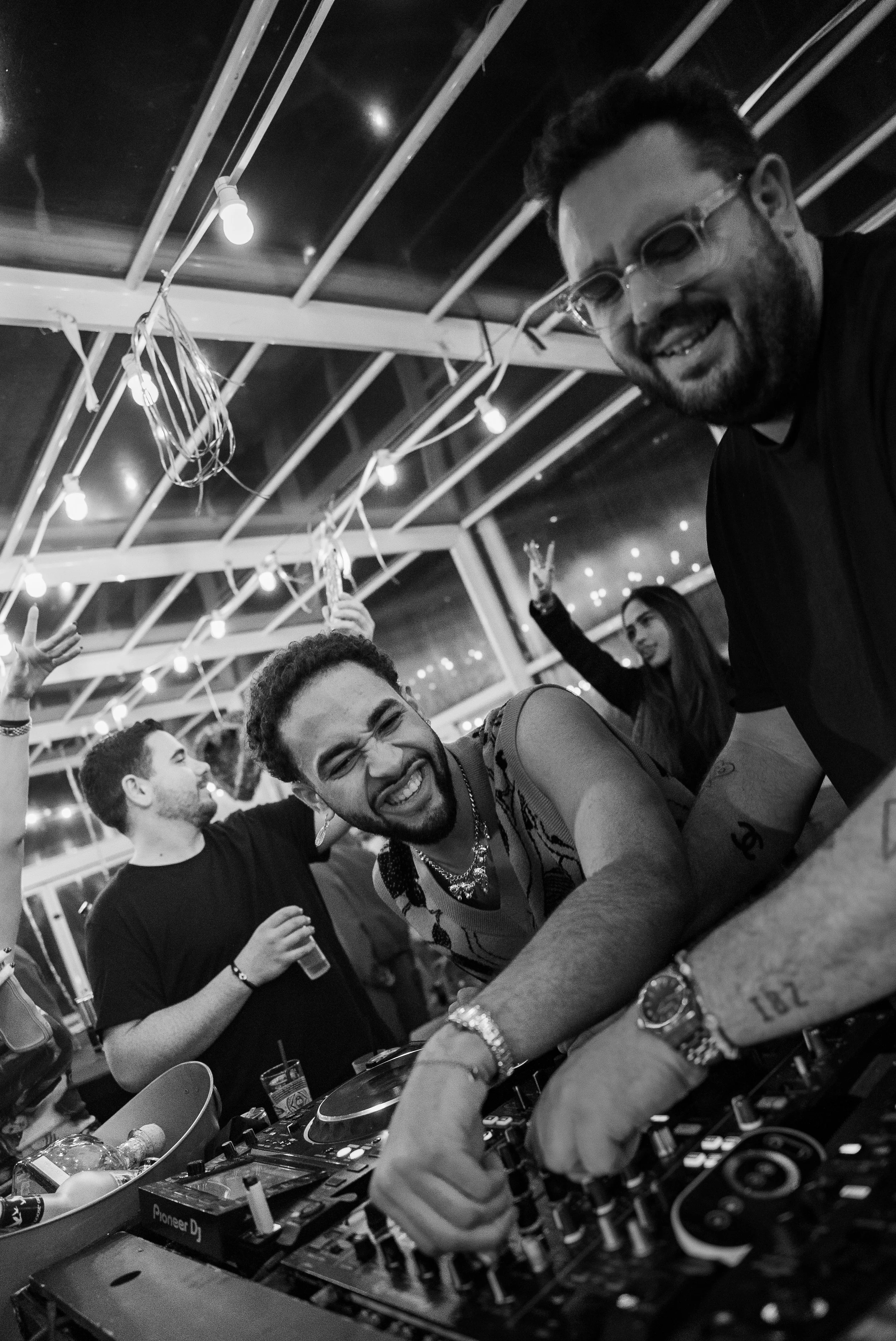 Groupe de personnes dansant et souriant dans une fête, avec un DJ à l'avant, dans un lieu éclairé par des lumières suspendues