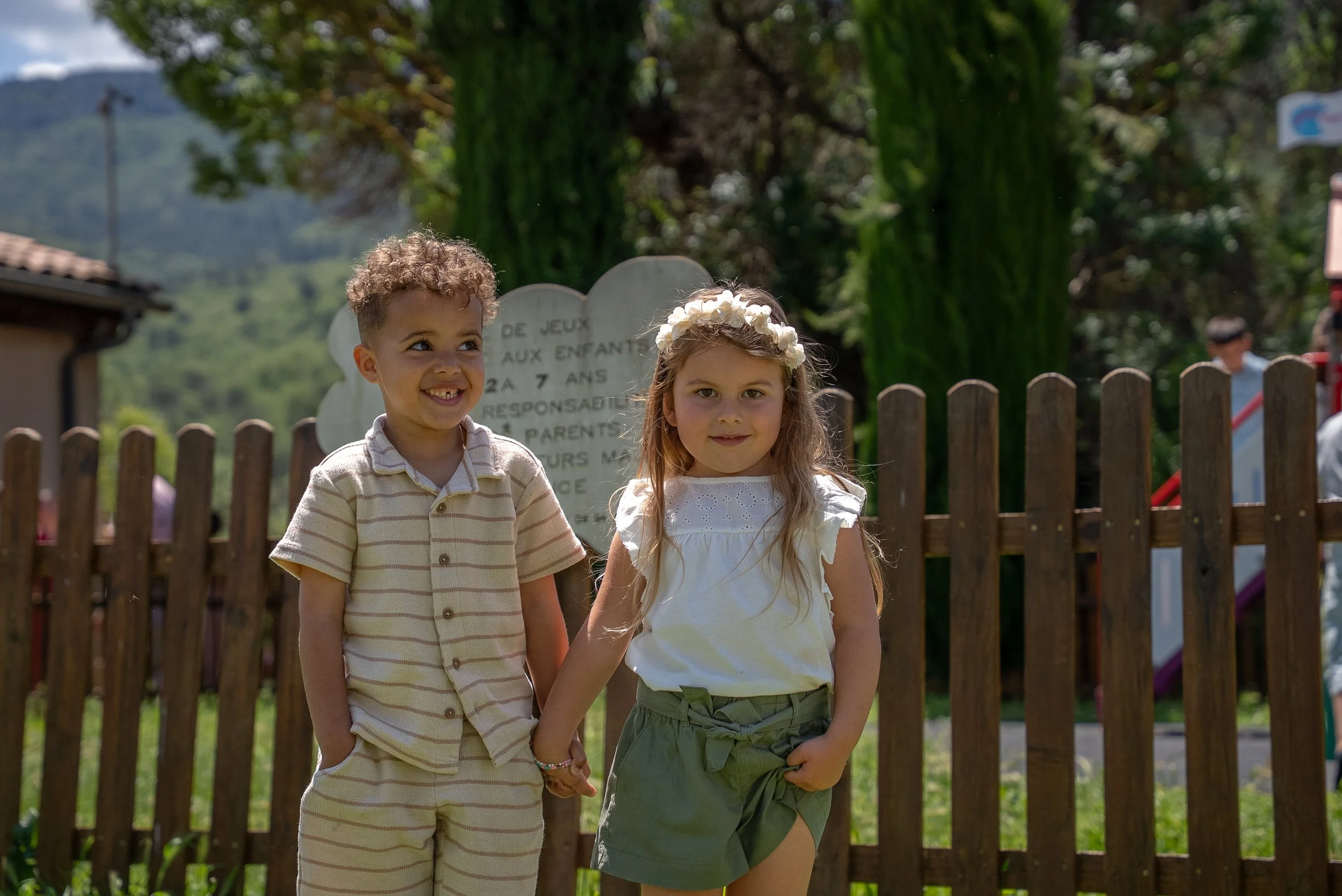 Deux enfants, un garçon et une fille, tenant la main, souriant à l'extérieur devant une clôture en bois, avec des arbres et un panneau en arrière-plan.