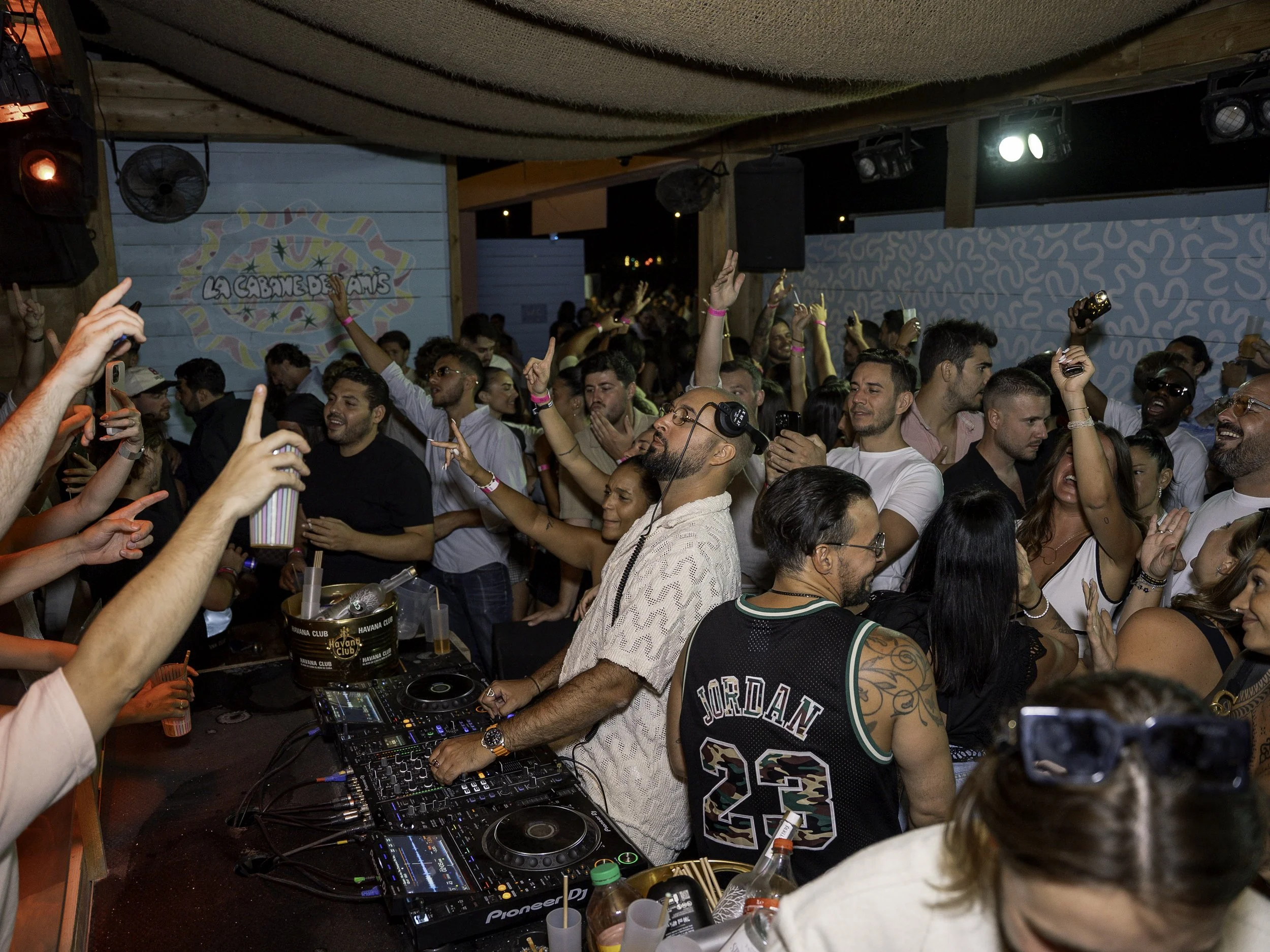 Une foule de personnes dans une fête, certaines dansent, d'autres prennent des photos ou regardent la scène, avec un DJ au centre jouant de la musique, ambiance festive et animée.