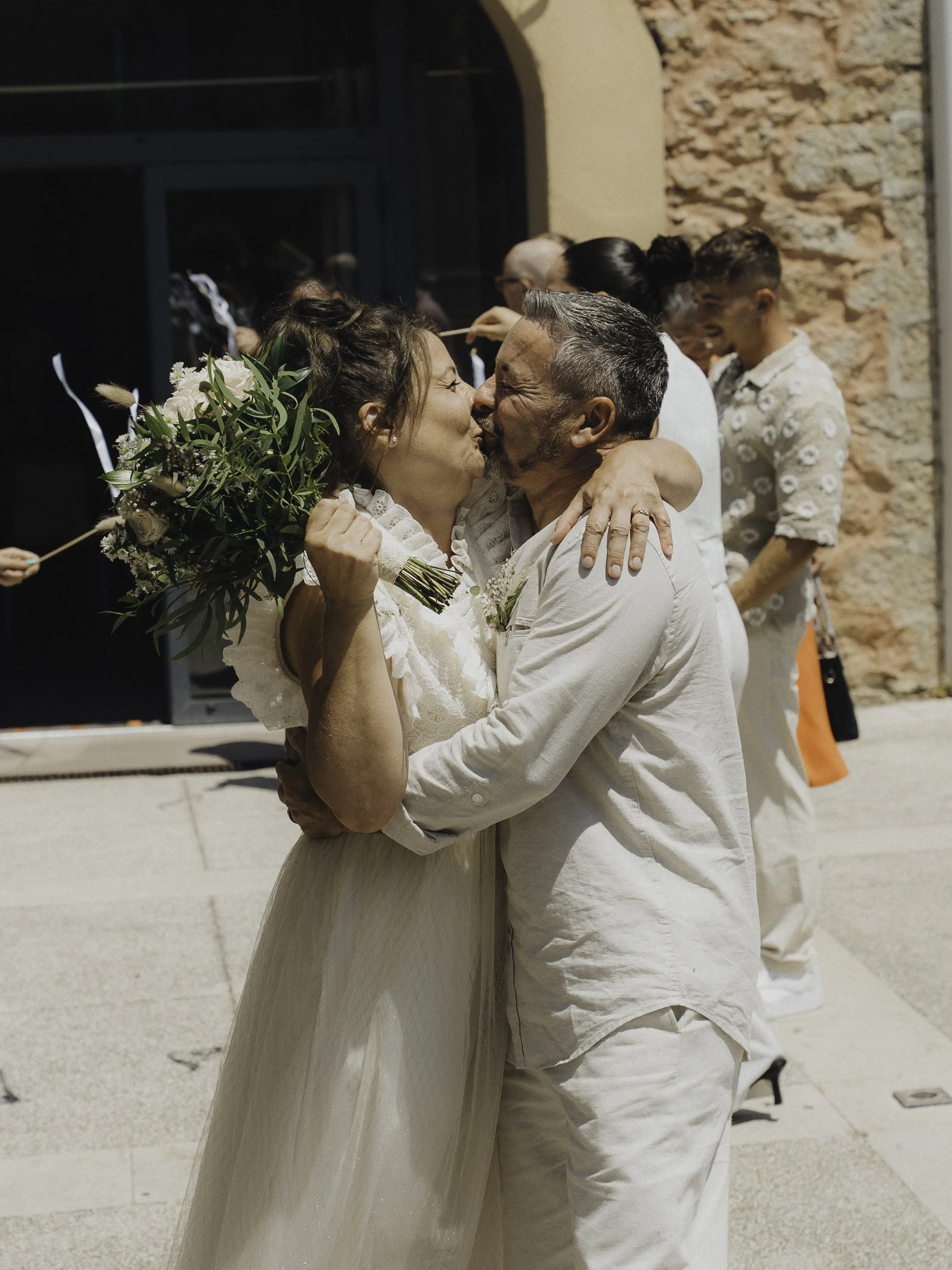 Un couple en mariage s'embrasse avec beaucoup d'émotion, entouré de proches, en plein air devant une bâtisse en pierre.