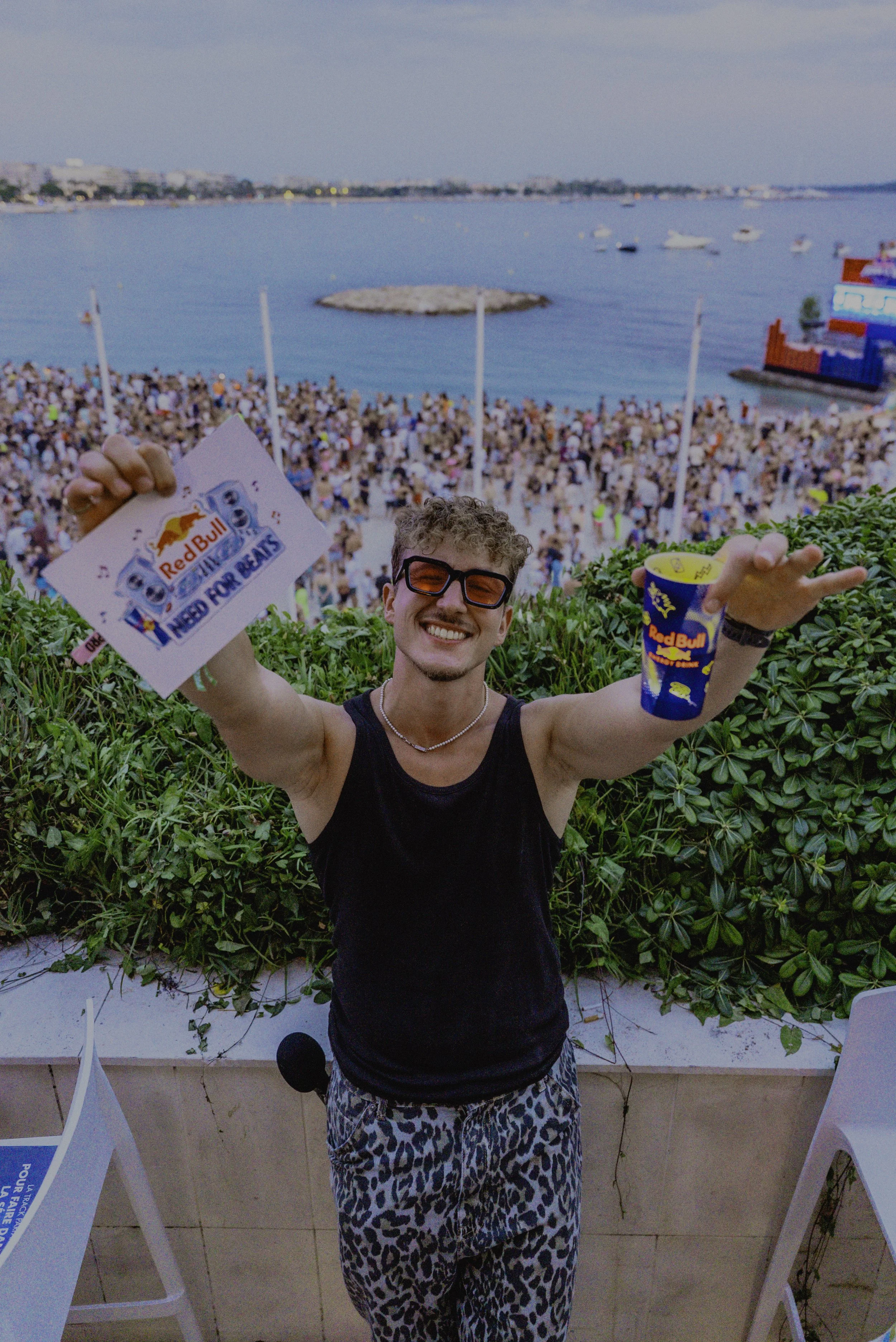 Un jeune homme souriant portant des lunettes de soleil, un t-shirt noir et un pantalon à motif léopard, tenant une tasse de Red Bull en plastique et un signe promotionnel de Red Bull, avec une foule de personnes sur la plage et la mer en arrière-plan