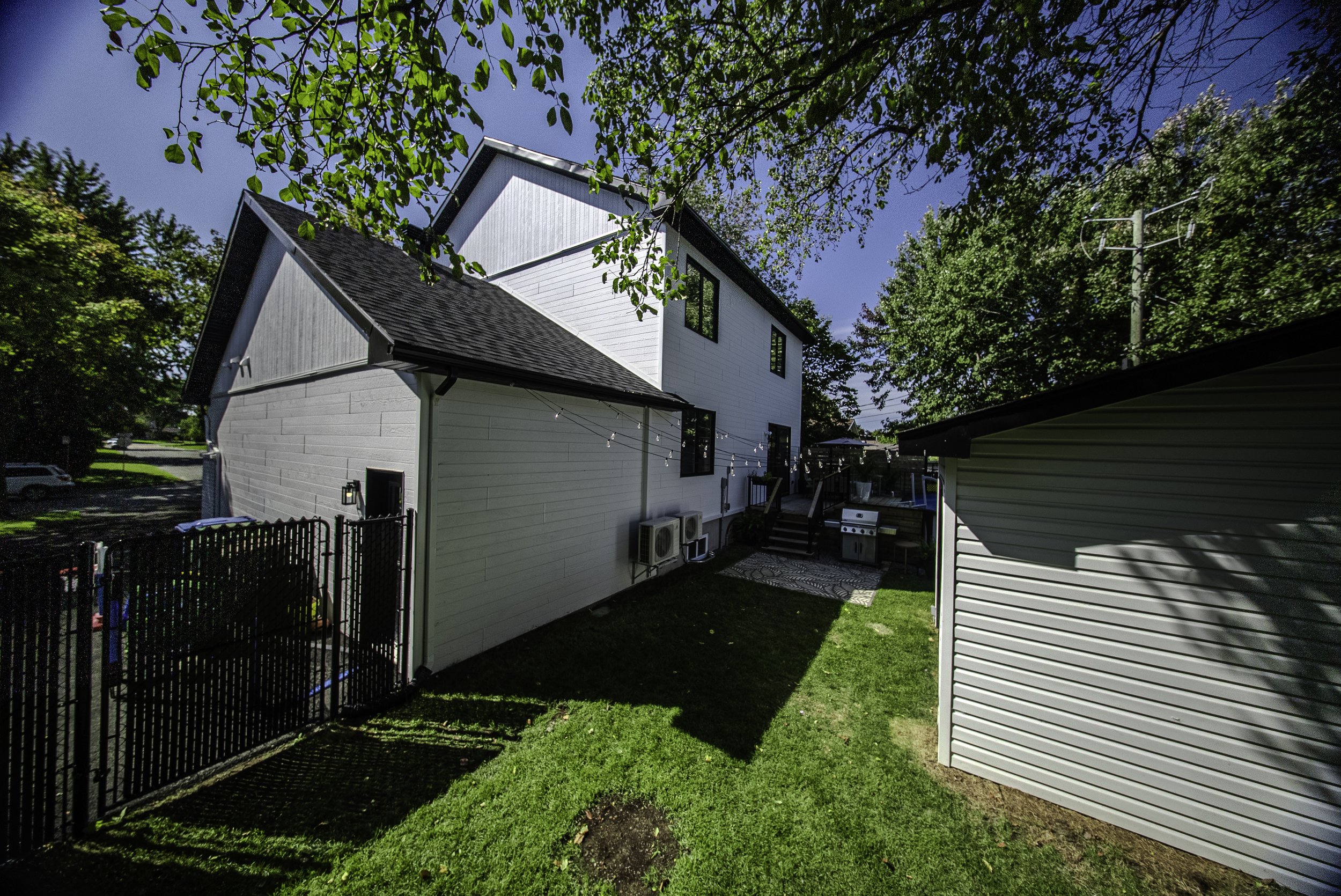 Vue d'une maison moderne blanche avec un étage, un toit en pente, un espace extérieur avec barbecue et un patio, entourée de verdure et de grands arbres, sous un ciel clair.