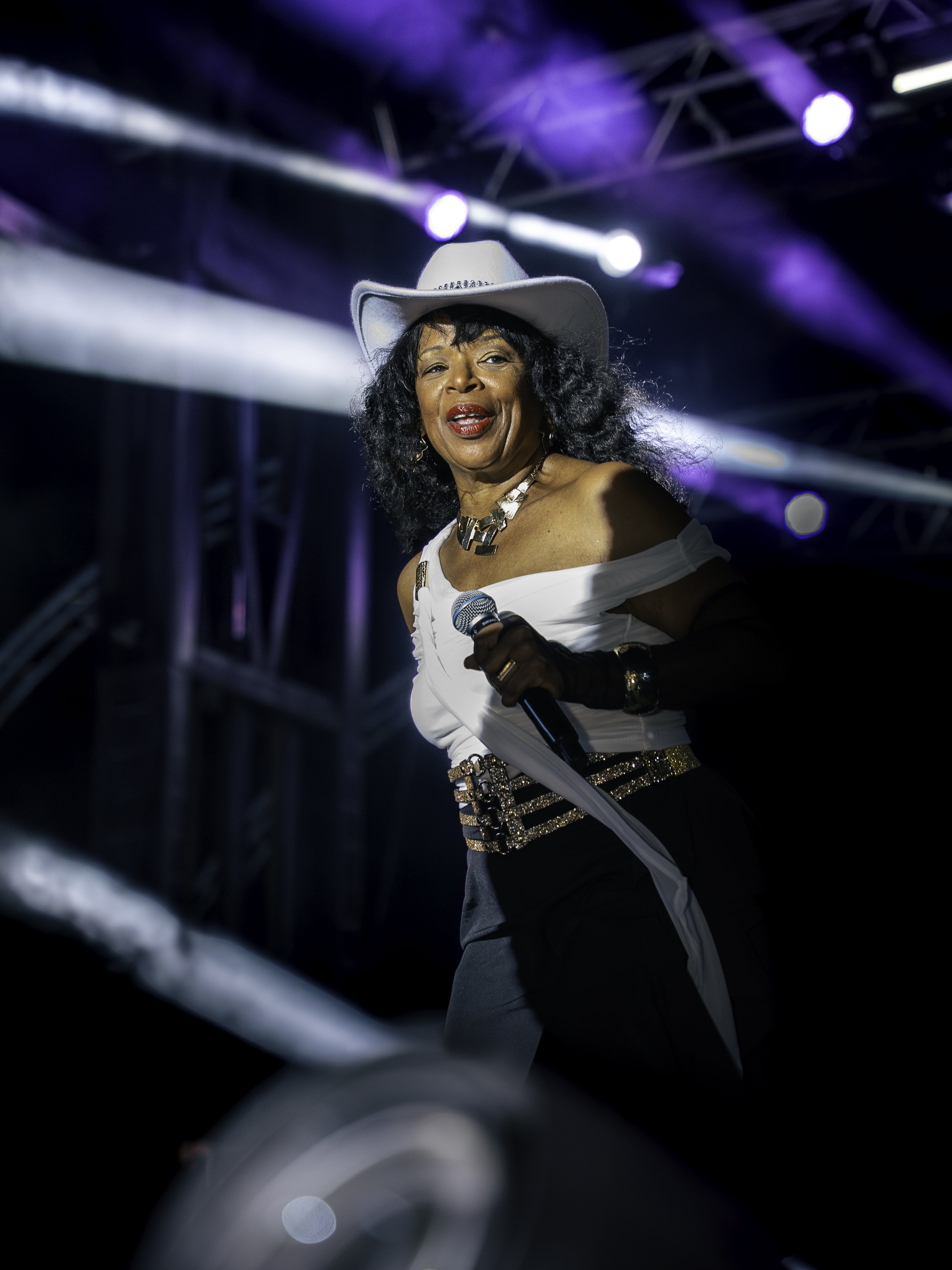 Femme chanteuse sur scène avec un chapeau blanc, holding un micro, sous des lumières violettes et blanches.