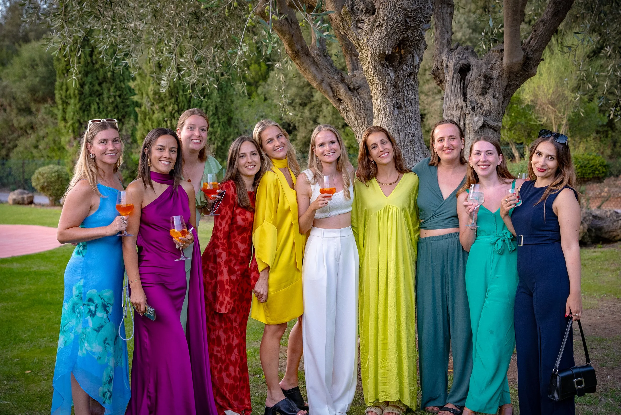 Groupe de femmes joyeuses en tenue colorée, tenant des verres de boisson, posant devant un arbre dans un jardin.
