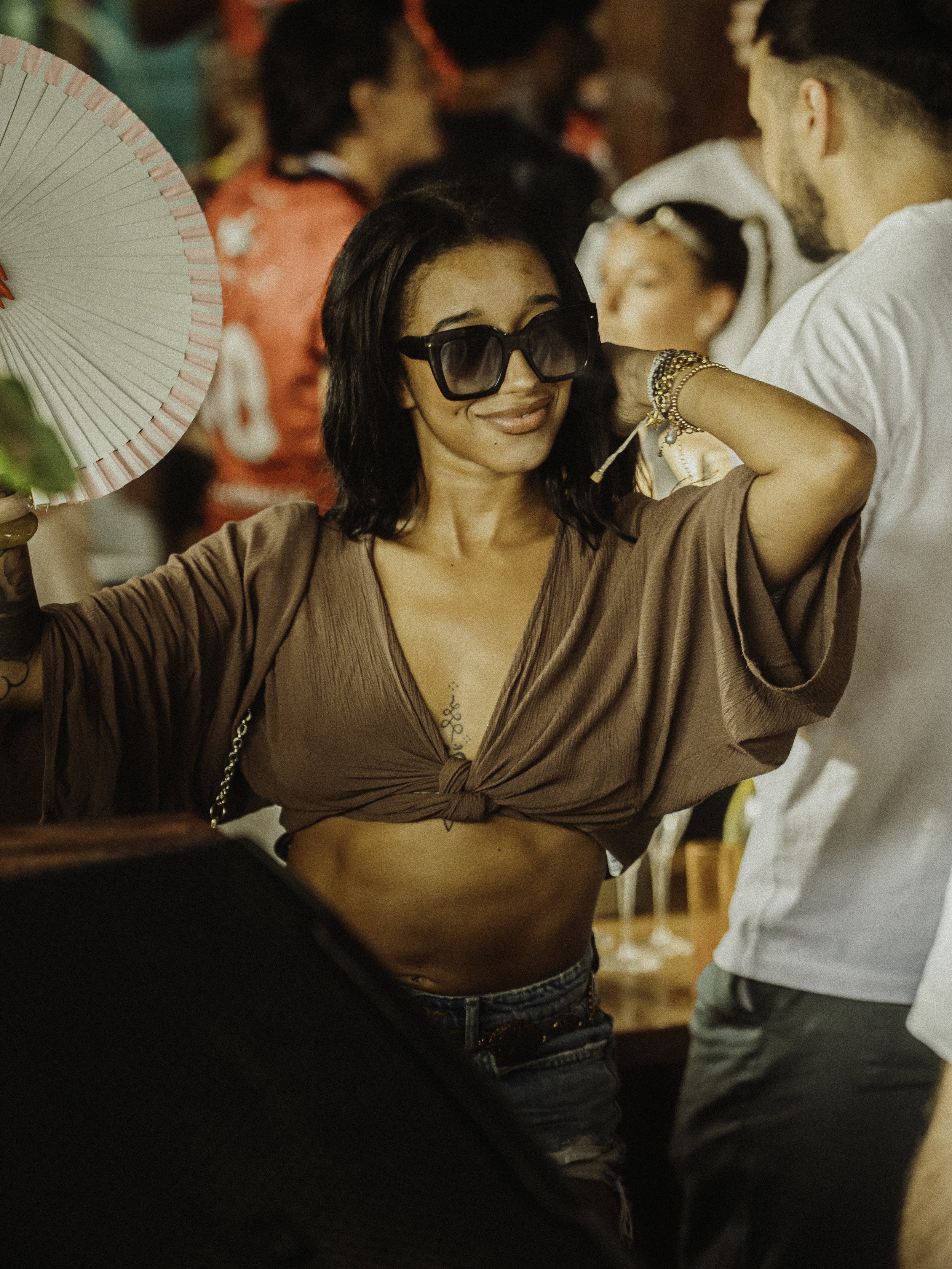 Femme souriante portant des lunettes de soleil noires, t-shirt beige noué à la taille, en train de danser ou de faire un mouvement dans une fête ou un lieu de rassemblement