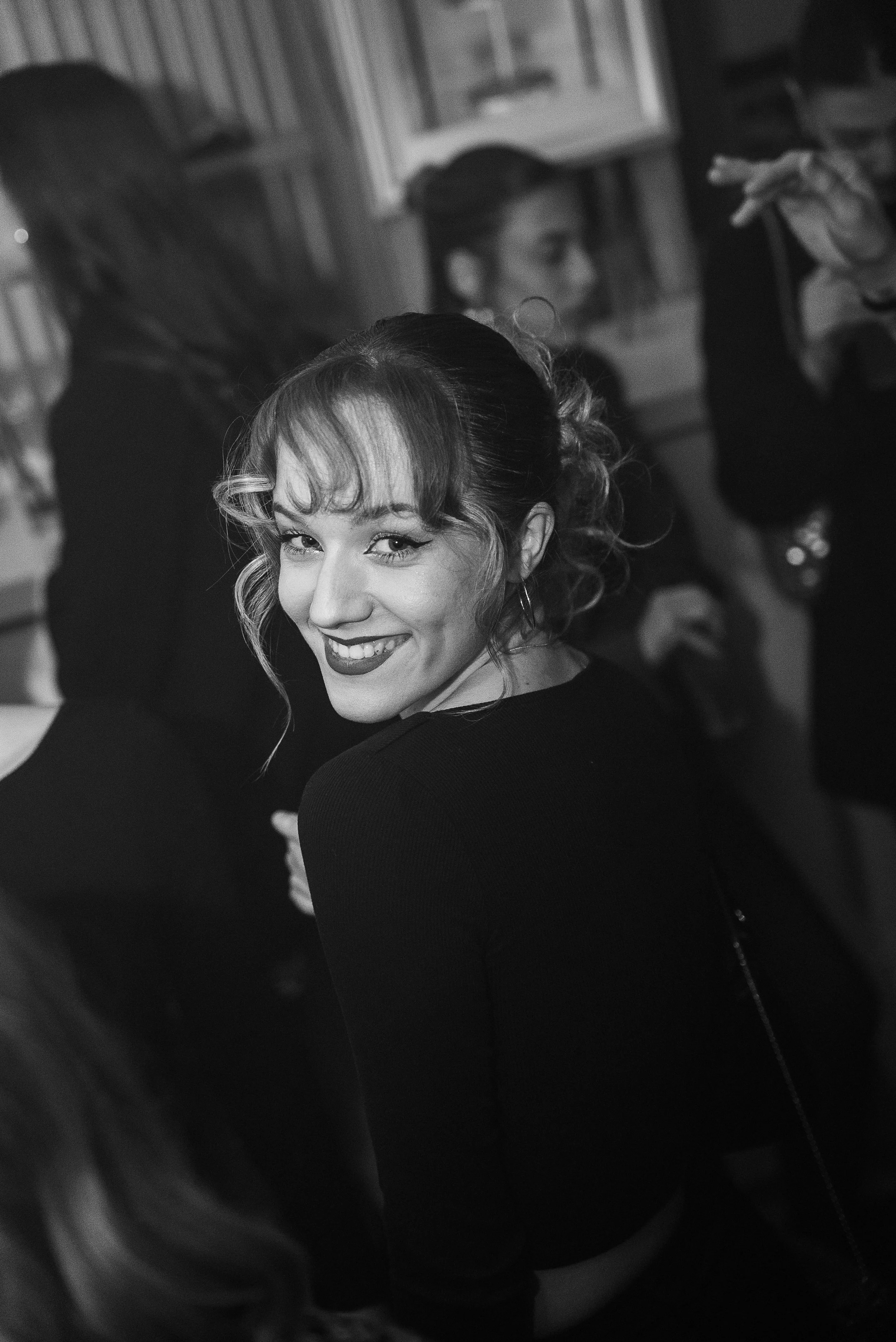 Une femme souriante avec des boucles d'oreilles et des cheveux attachés, entourée d'autres personnes dans un environnement intérieur, en noir et blanc.