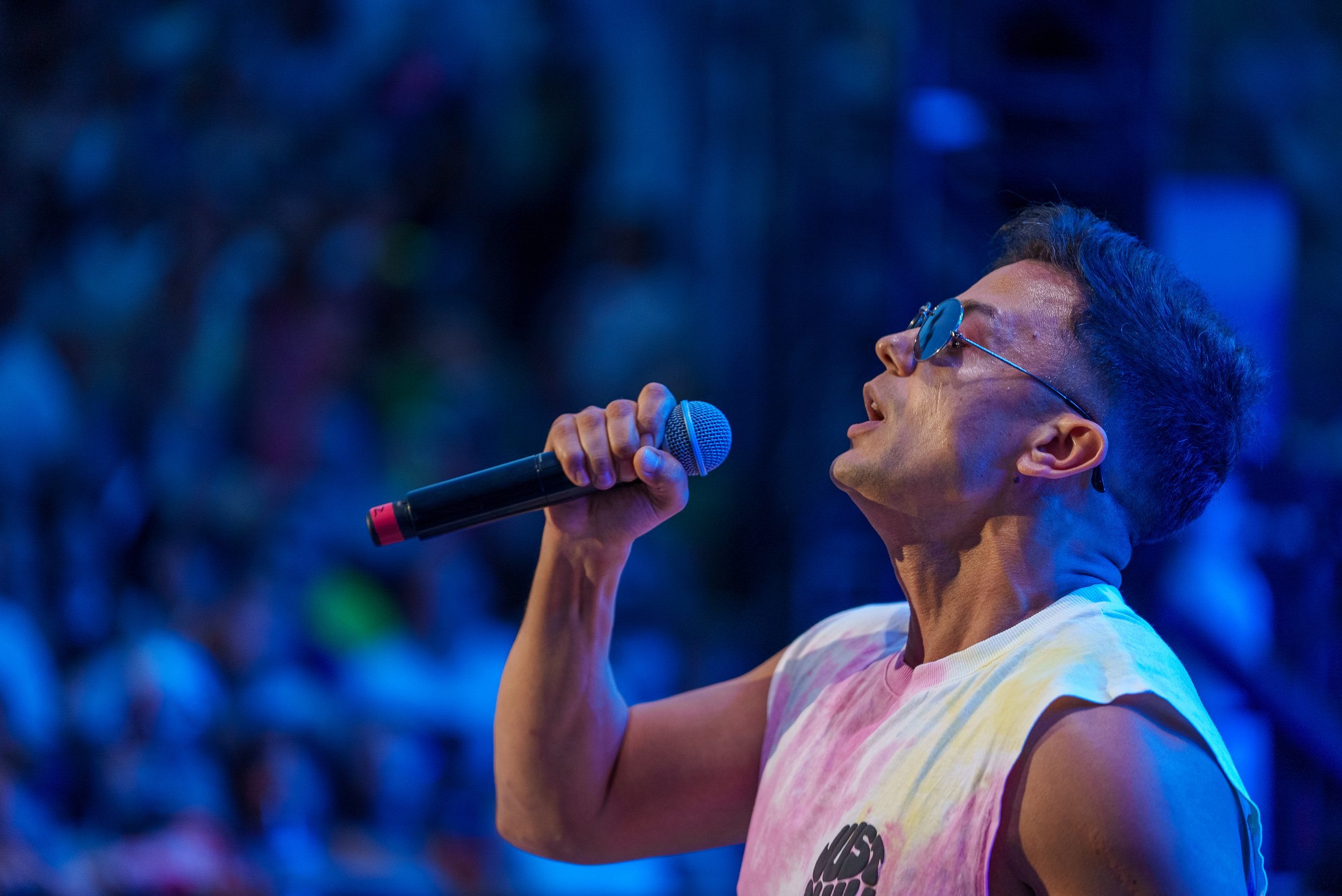 Un homme chantant avec un microphone, portant des lunettes de soleil, contre un fond flou de couleurs sombres et bleues.