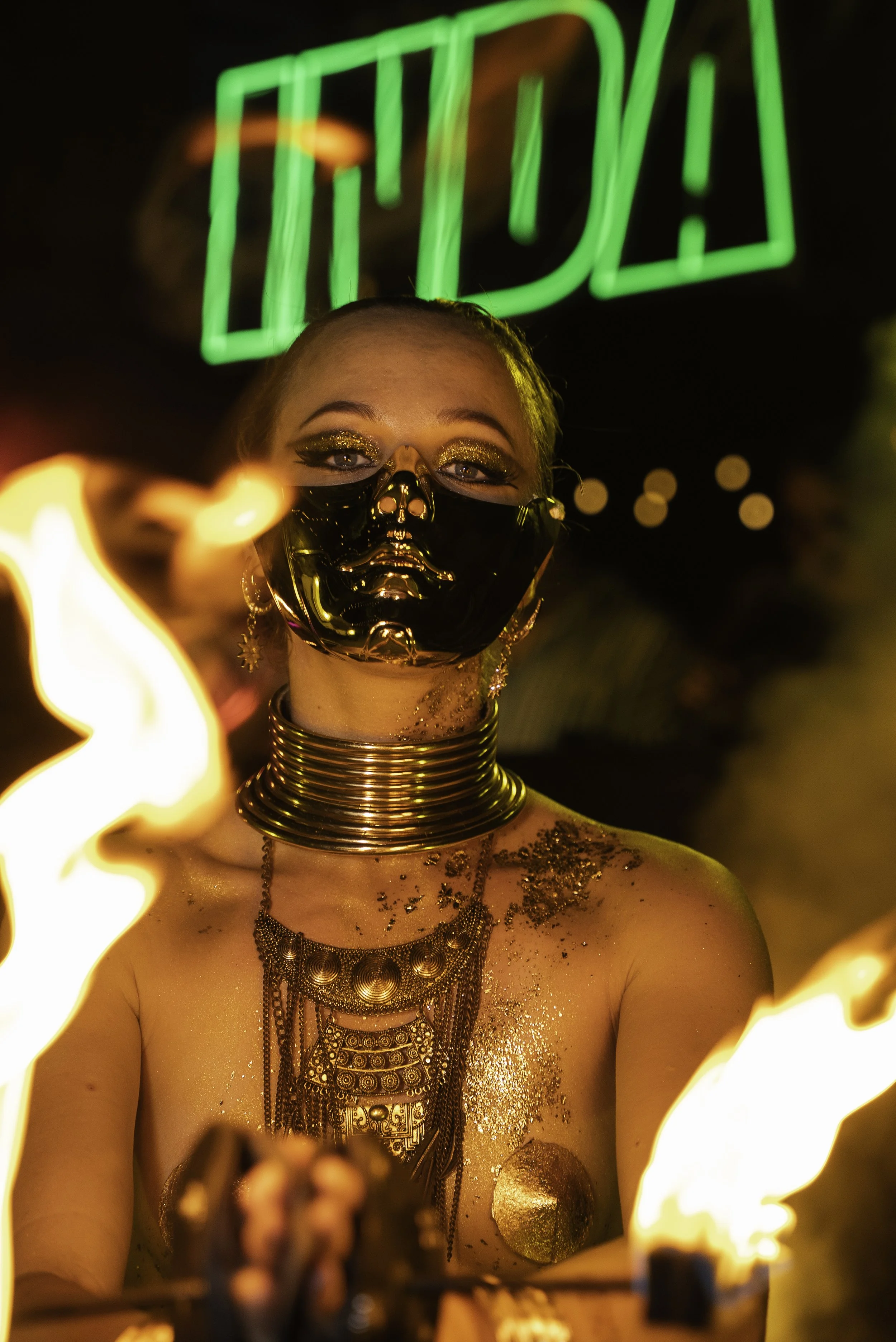 Une femme portant un masque noir brillant avec un crâne en or, des bijoux en or et des paillettes dorées, entourée de flammes, avec un néon vert en arrière-plan. Elle semble participer à une performance artistique.