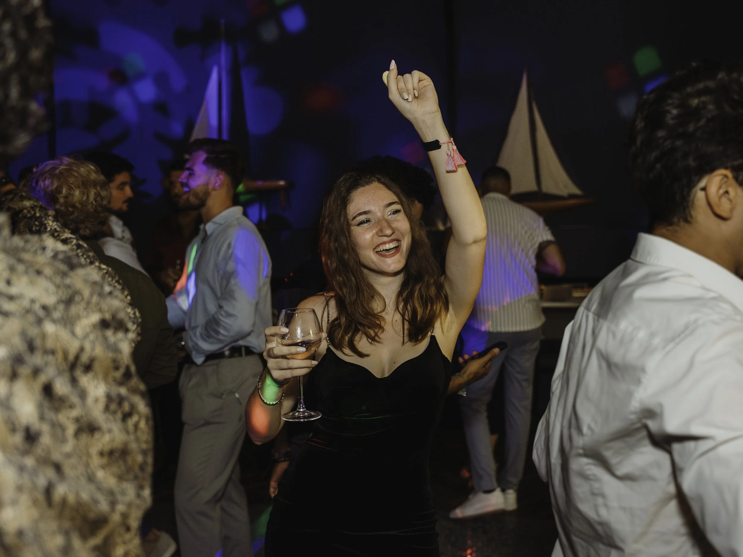 Femme joyeuse en robe noire, levant une main en l'air, tenant un verre de vin dans l'autre, dans une fête ou une discothèque avec d'autres personnes