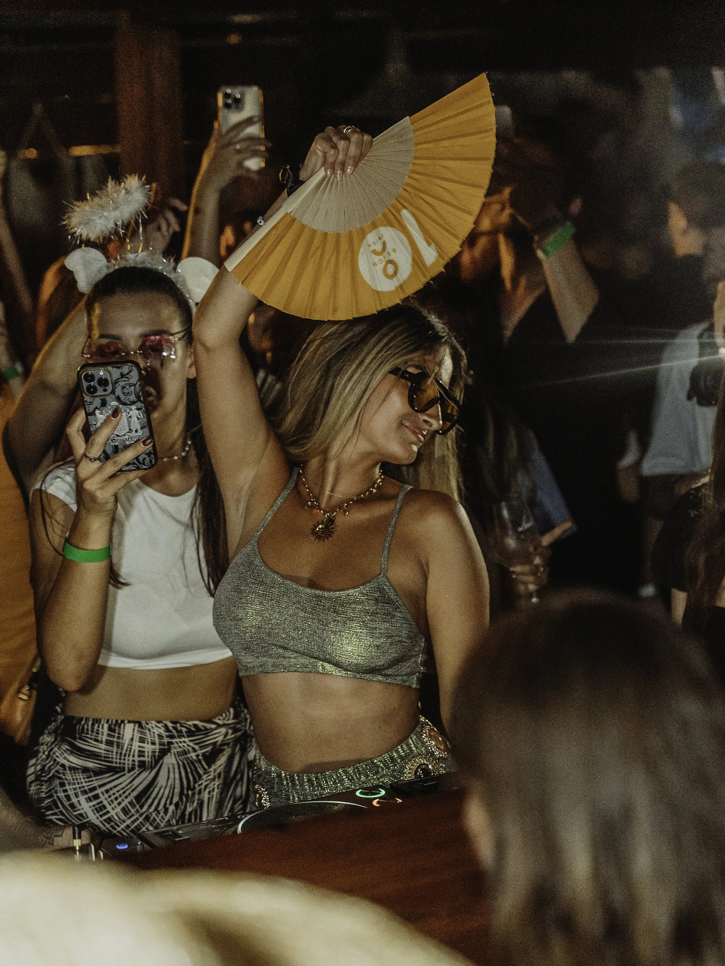 Une femme danseuse portant un haut doré et un pantalon à motif noir et blanc, tenant un éventail orange. Elle porte des lunettes de soleil et un collier orné. Autour d'elle, d'autres personnes prennent des photos lors d'une fête ou d'un concert, avec