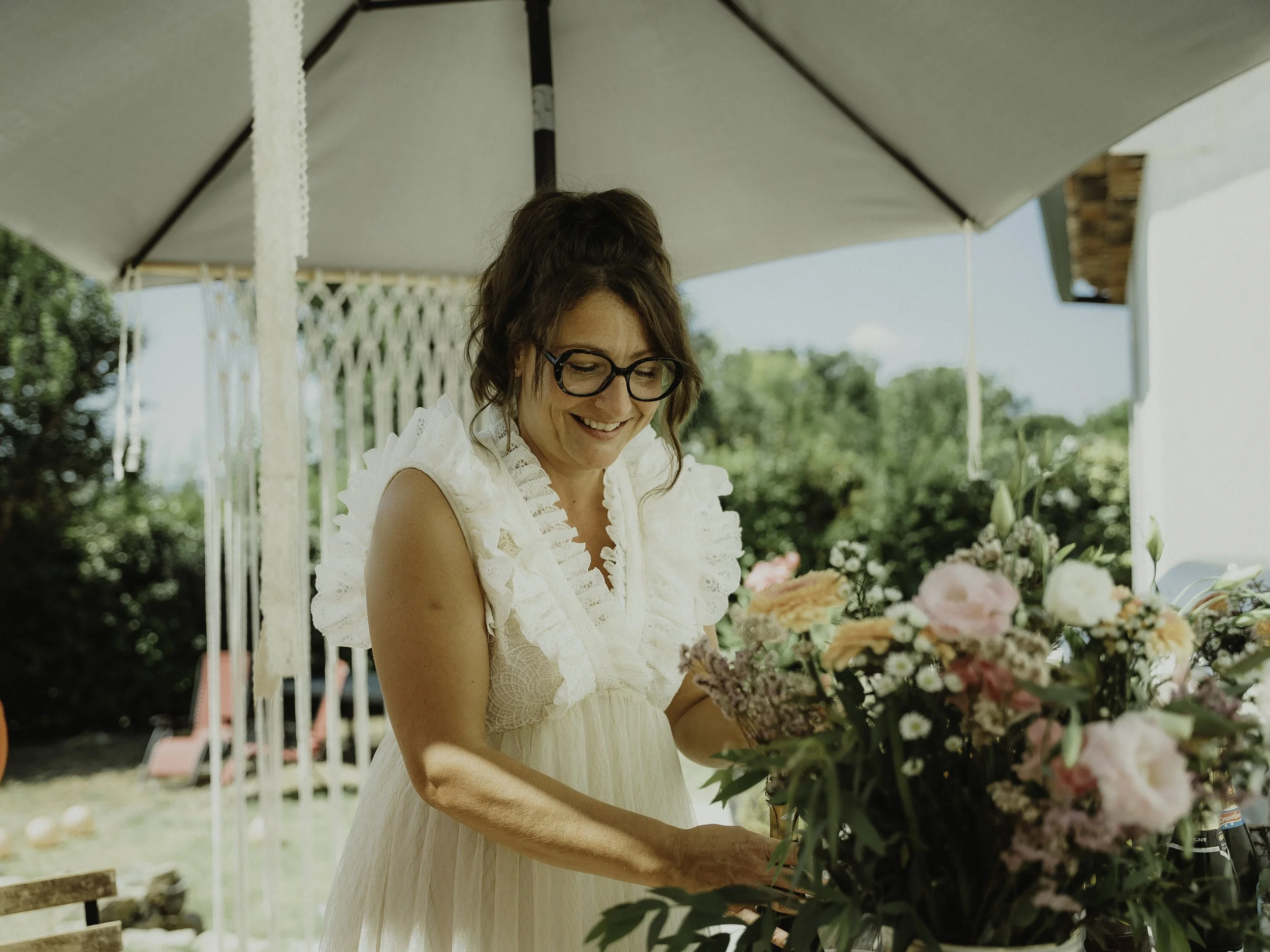 Une femme souriante, portant des lunettes, habillée en blanc, en train de faire un arrangement floral sous un parasol lors d'une journée ensoleillée.