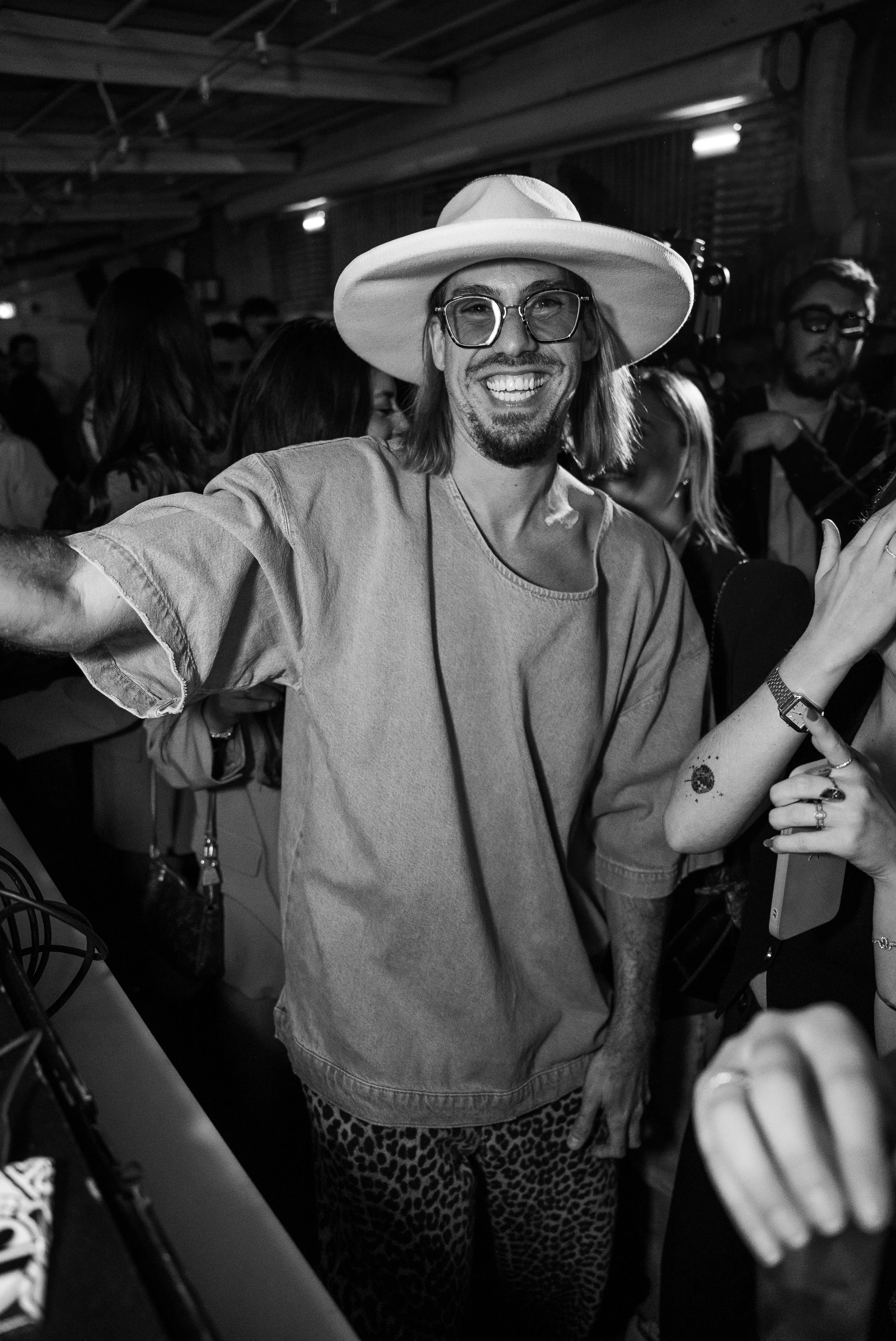 Un homme souriant portant un grand chapeau blanc, des lunettes rondes, un t-shirt ample et des pantalons à motifs léopard, entouré de plusieurs personnes dans un lieu animé.