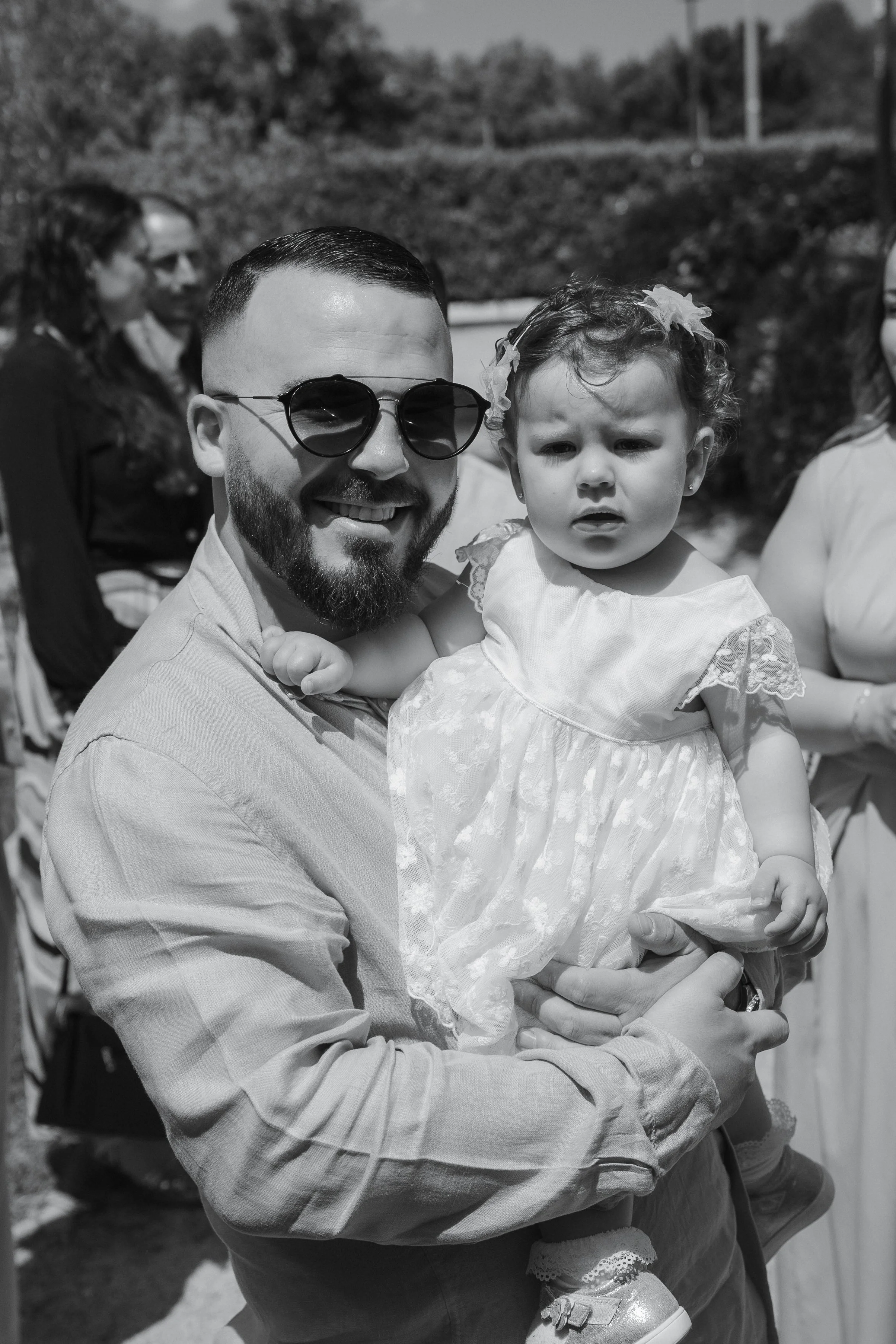 Un homme souriant portant des lunettes de soleil, tenant une petite fille dans ses bras lors d'un événement en plein air.