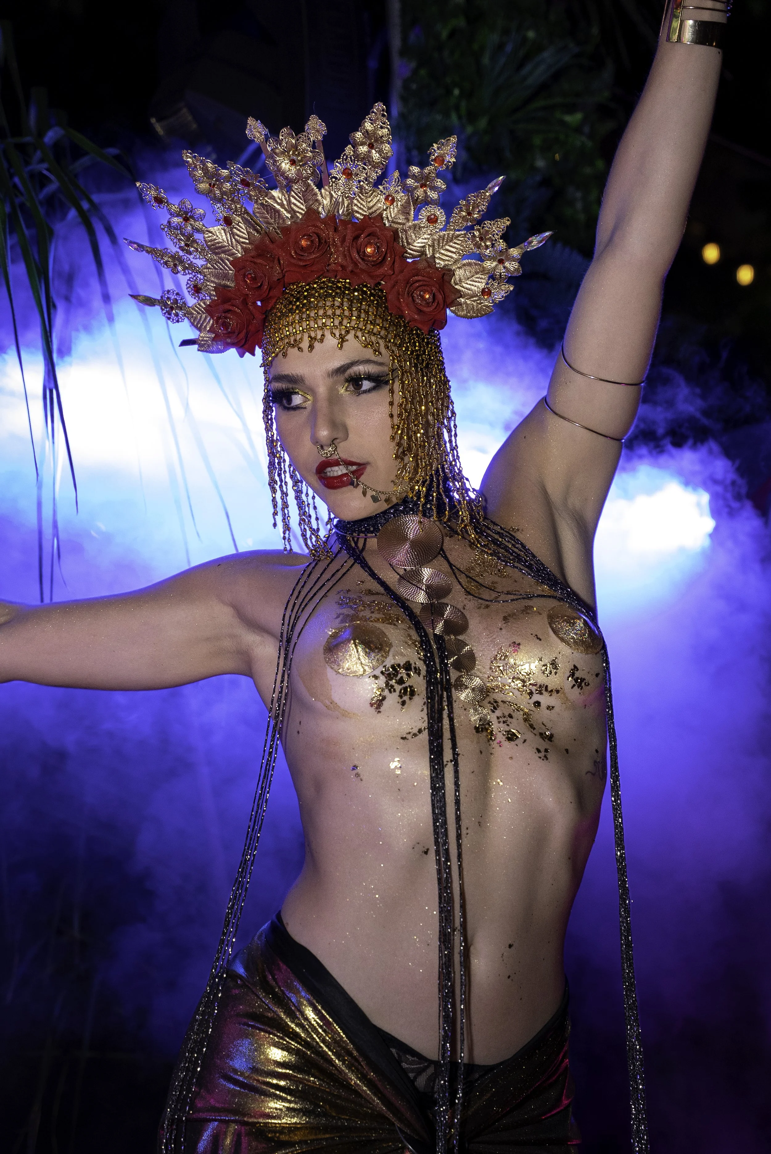 Une femme portant une coiffe orné de roses rouges, de feuilles dorées et de bijoux, avec des vêtements transparents et dorés, dans un décor coloré et mystérieux ambigu. Elle pose avec une expression expressive sur le visage, sous un éclairage coloré.