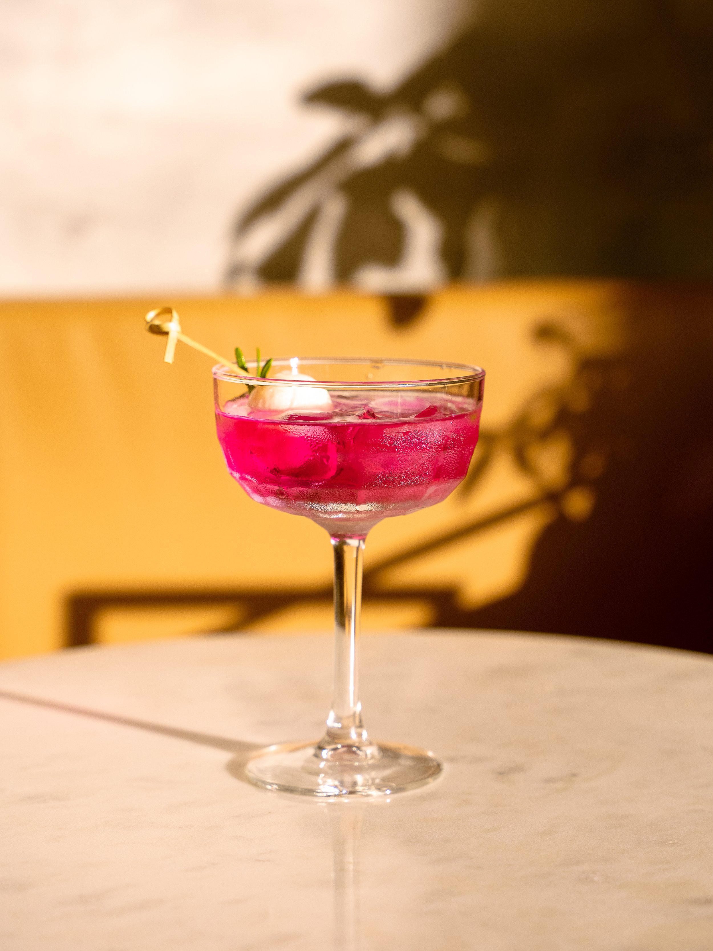 verre à cocktail rose avec une olive et une branche de romarin, posé sur une table en marbre