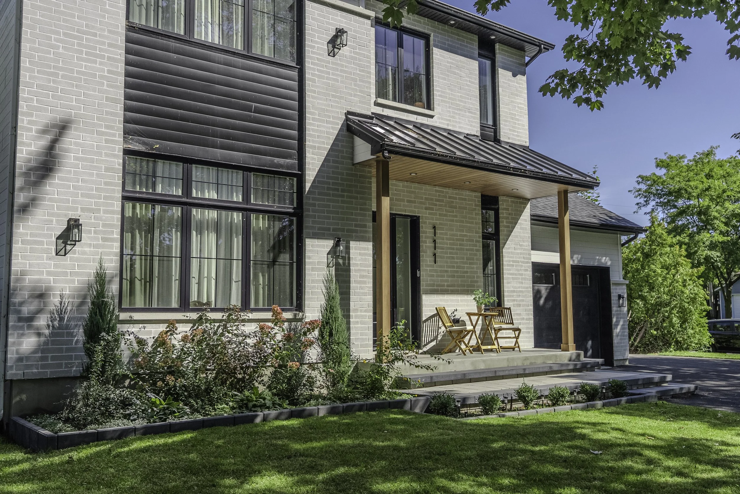 Façade d'une maison moderne avec un petit porche en bois et jardin avec plantes et arbustes.