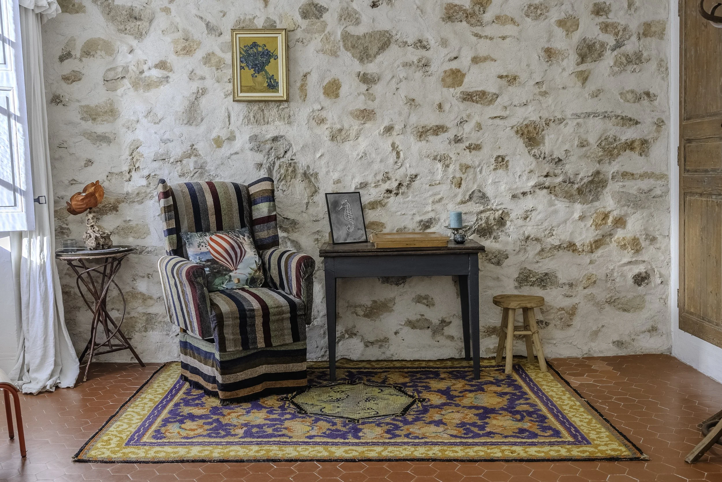 Salon avec fauteuil à rayures, table en bois avec cadre photo de cheval, petite lampe à côté, tapis coloré sur sol en carreaux de terre cuite, mur en pierre avec tableau et porte en bois.