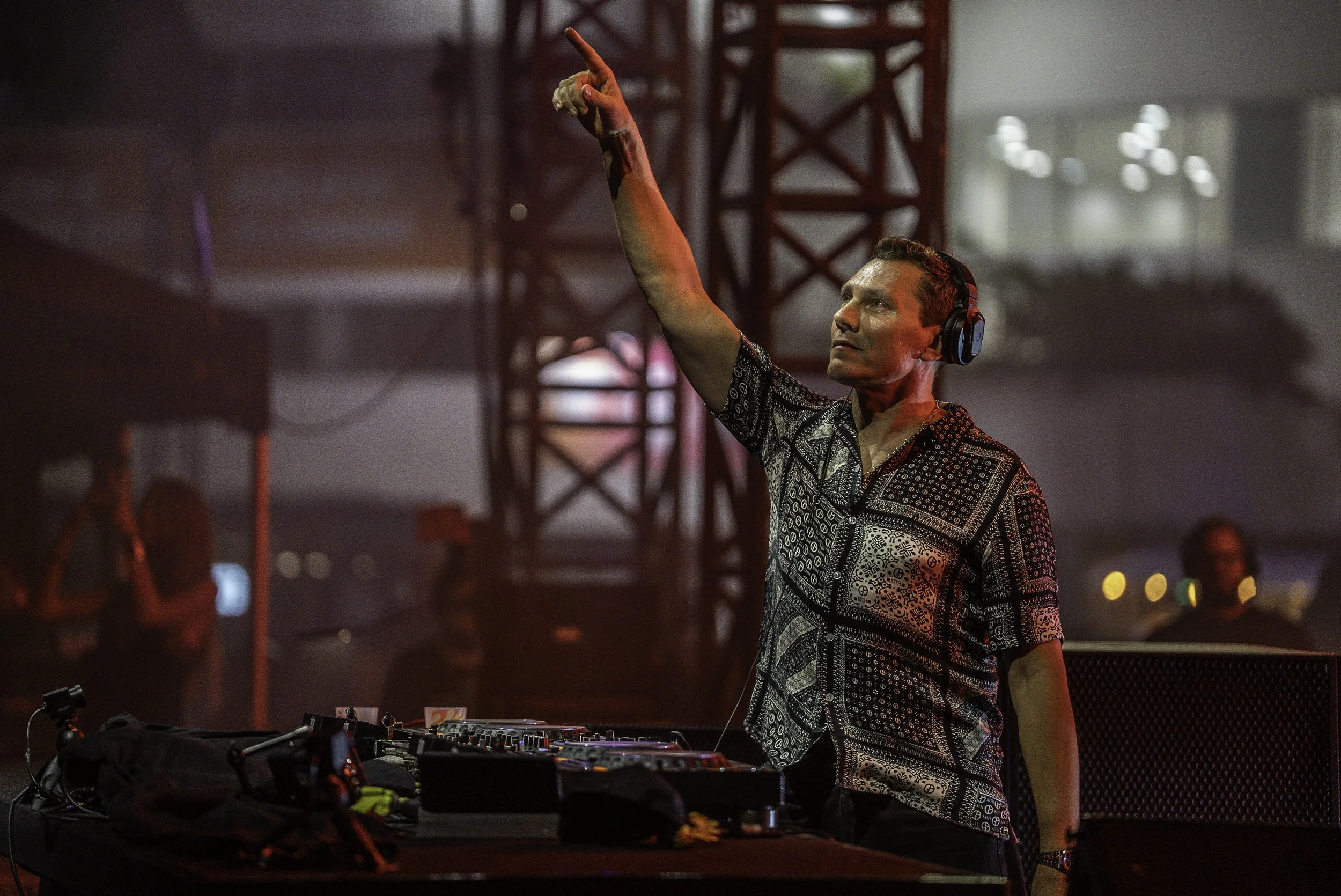 Un DJ portant un casque et un chemisier à motifs, utilisant un mixeur lors d'un événement en intérieur, avec des structures en métal en arrière-plan.