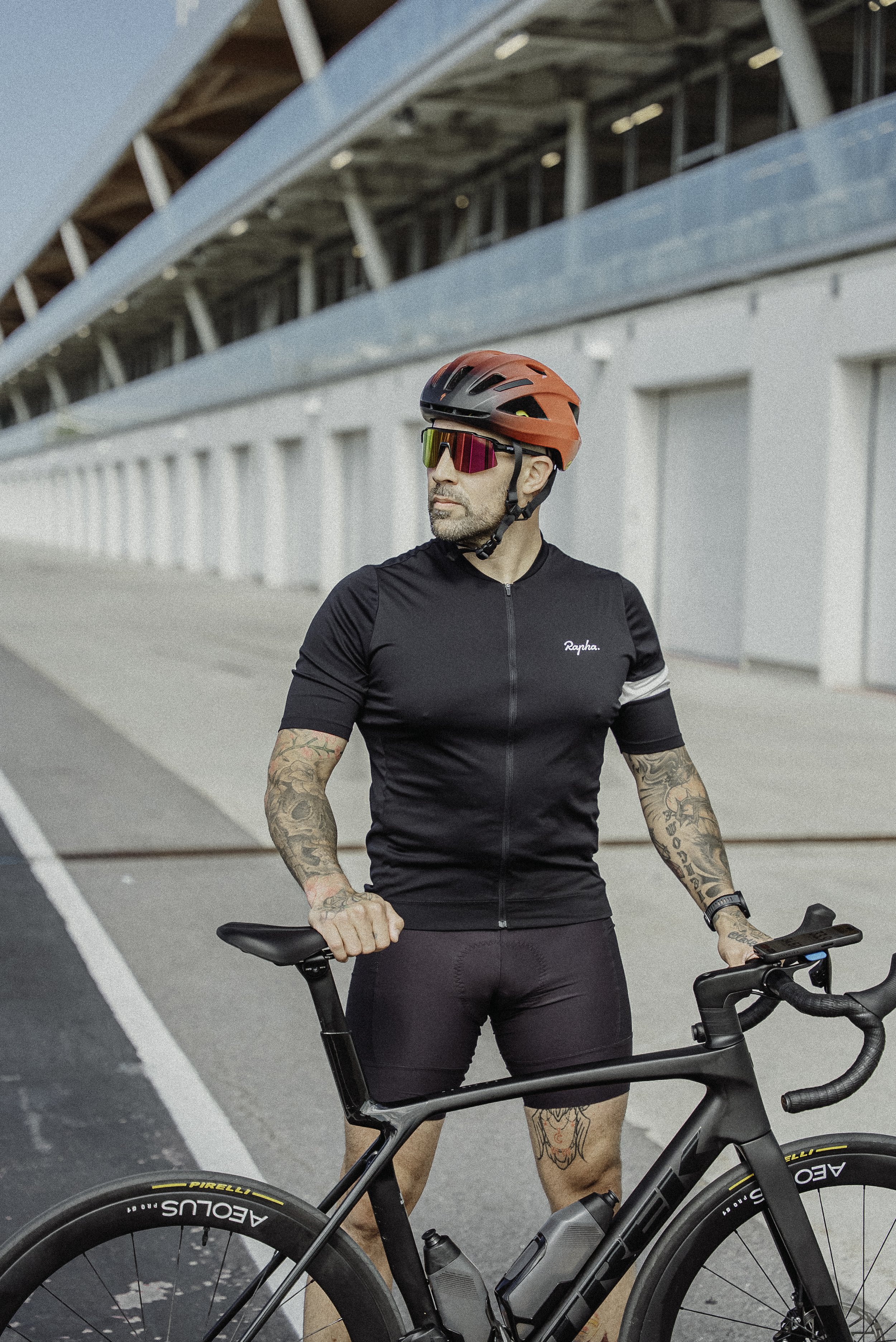 Un homme en tenue de cyclisme, portant un casque rouge et des lunettes de soleil, se tient près de son vélo dans un lieu urbain.