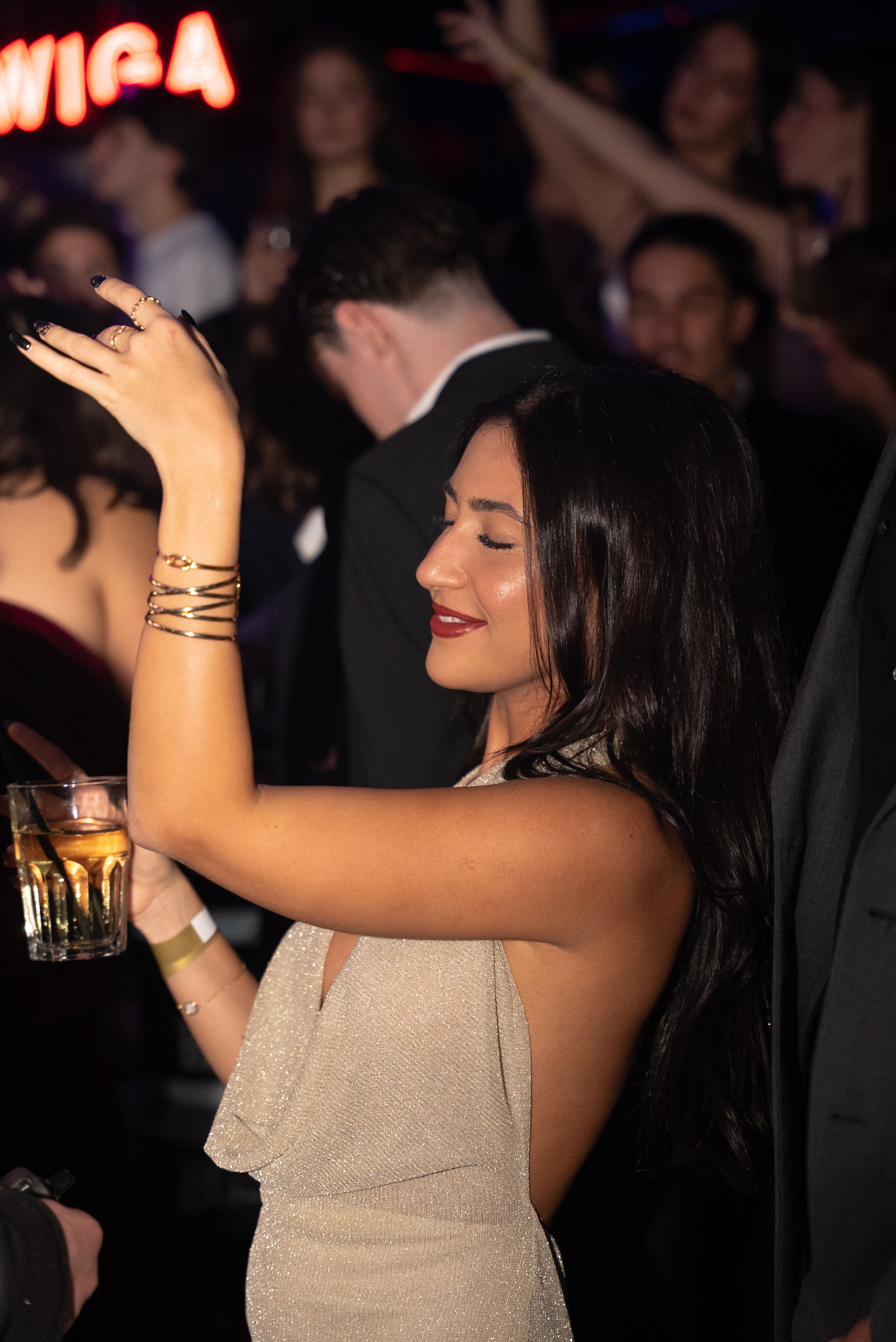 Une femme souriante avec des bijoux dorés et un cocktail dans une main, danse lors d'une fête ou d'une célébration en compagnie d'autres personnes chic dans un environnement festif.