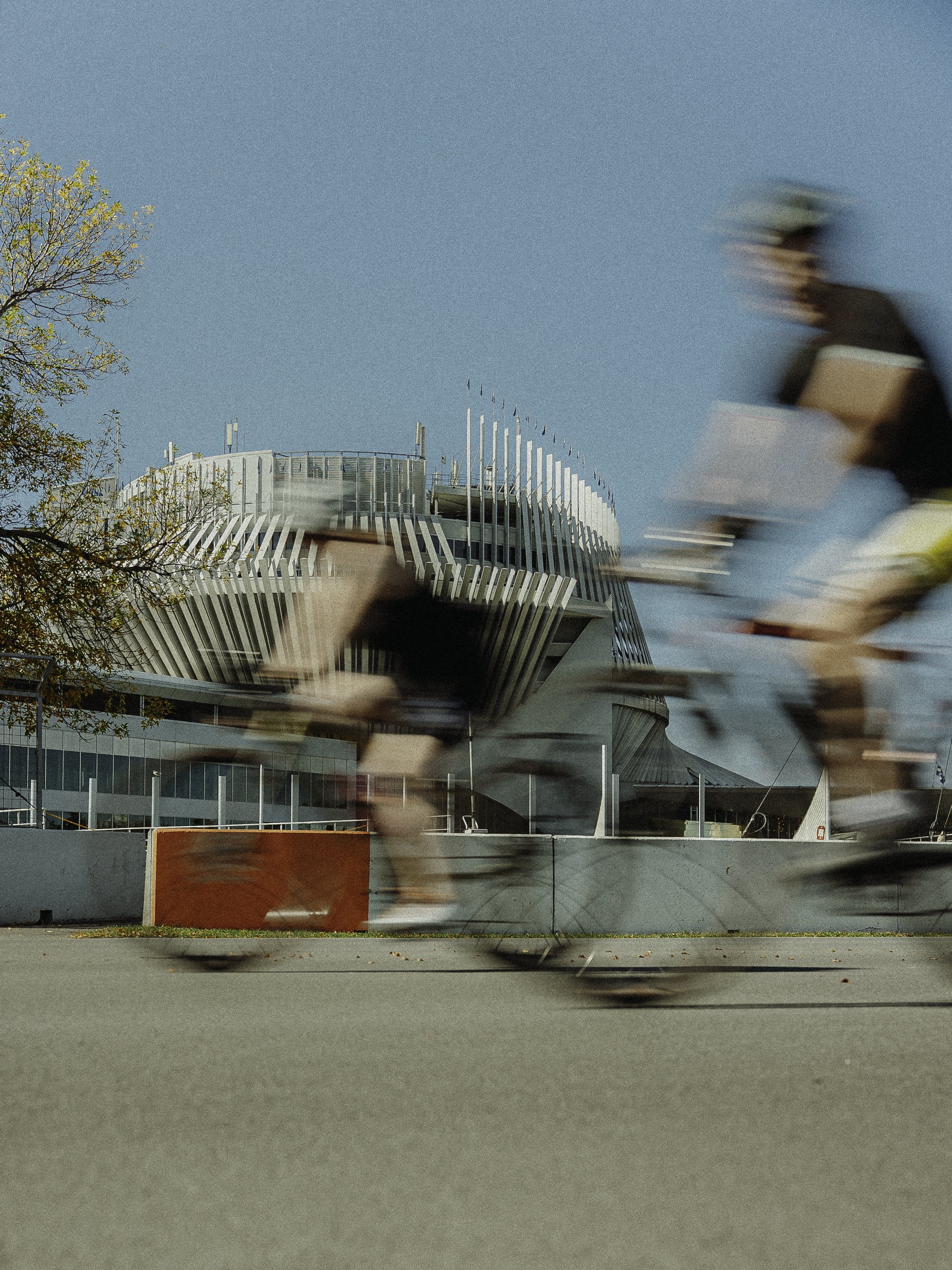 Deux cyclistes passent rapidement devant un bâtiment moderne avec une architecture distinctive, sous un ciel bleu.