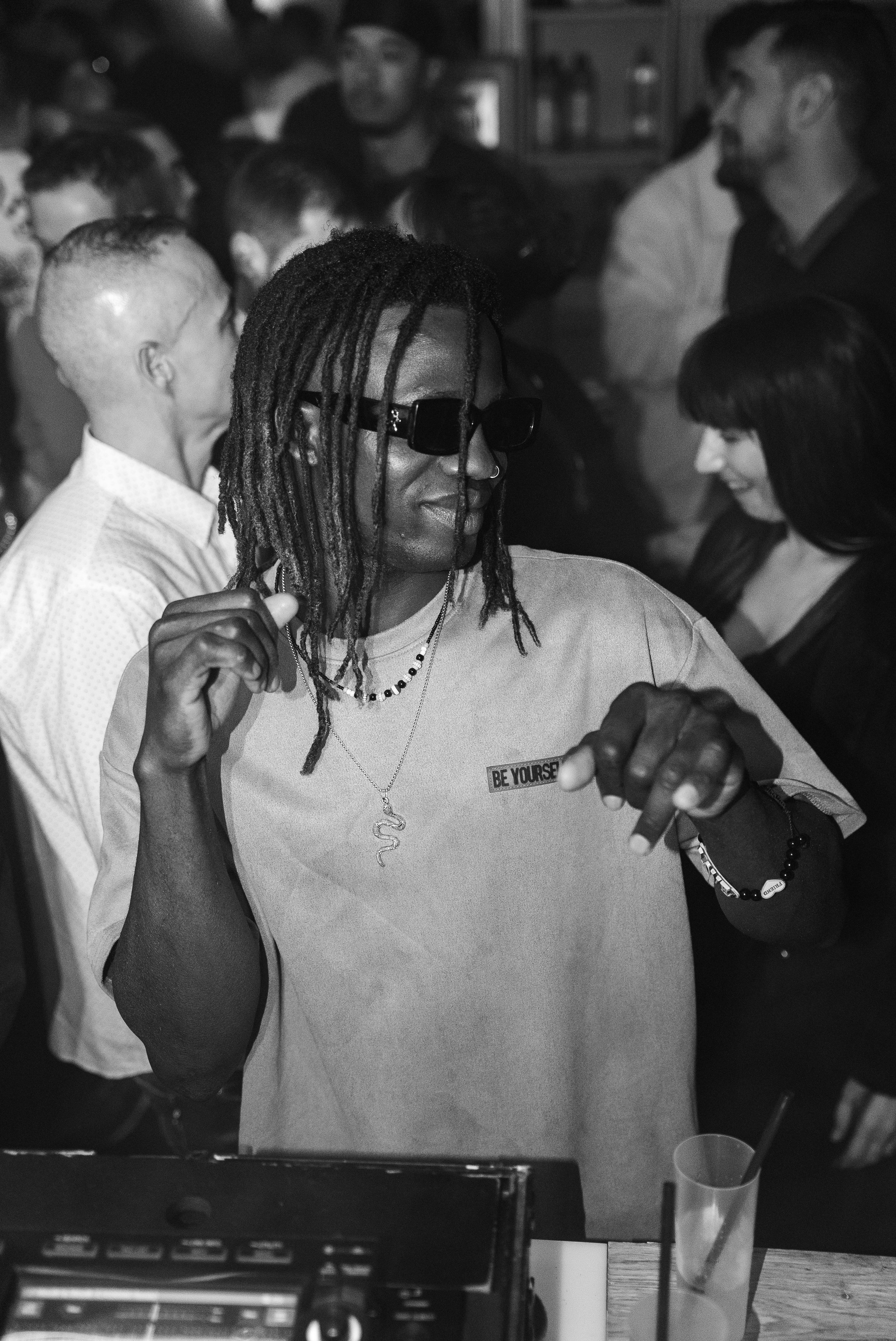 Homme avec dreadlocks portant des lunettes de soleil, une chemise avec la phrase 'BE YOURSELF', et des bijoux, en train de danser ou de faire un mouvement dans une fête ou un rassemblement, entouré d'autres personnes.