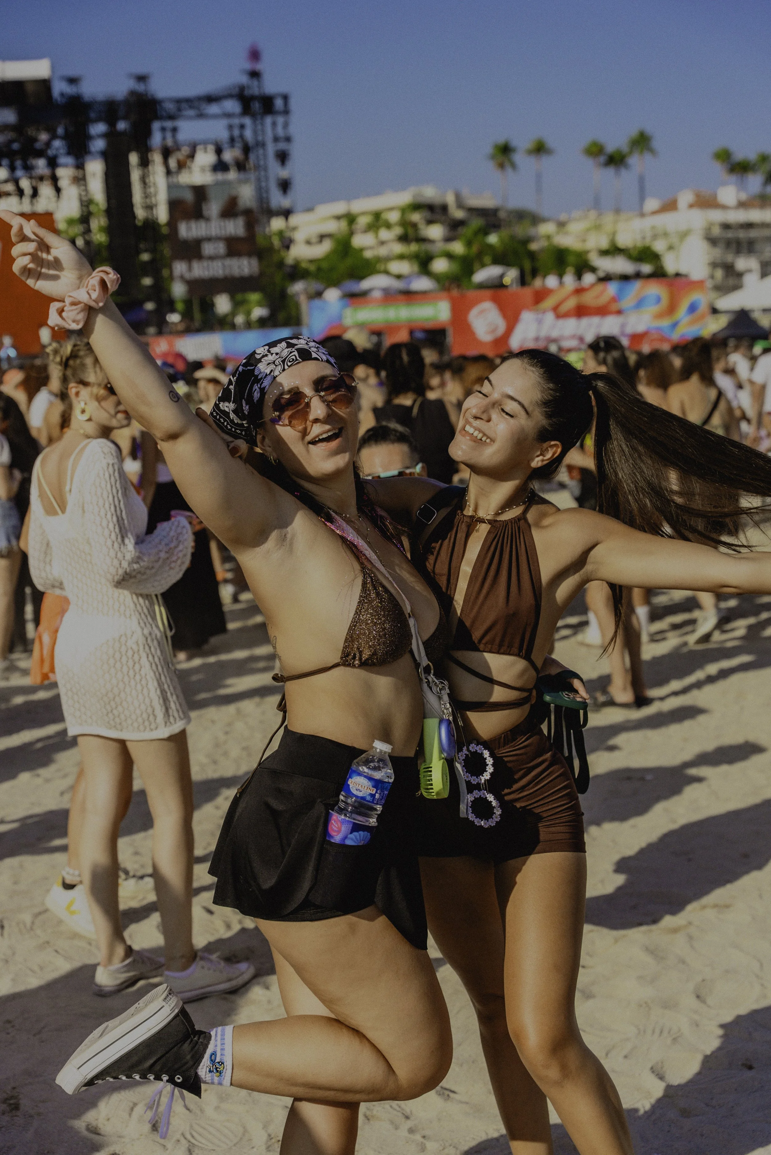 Deux femmes souriantes dansant sur la plage lors d'un festival en plein air, avec un public nombreux et des scènes en arrière-plan, sous un ciel bleu.