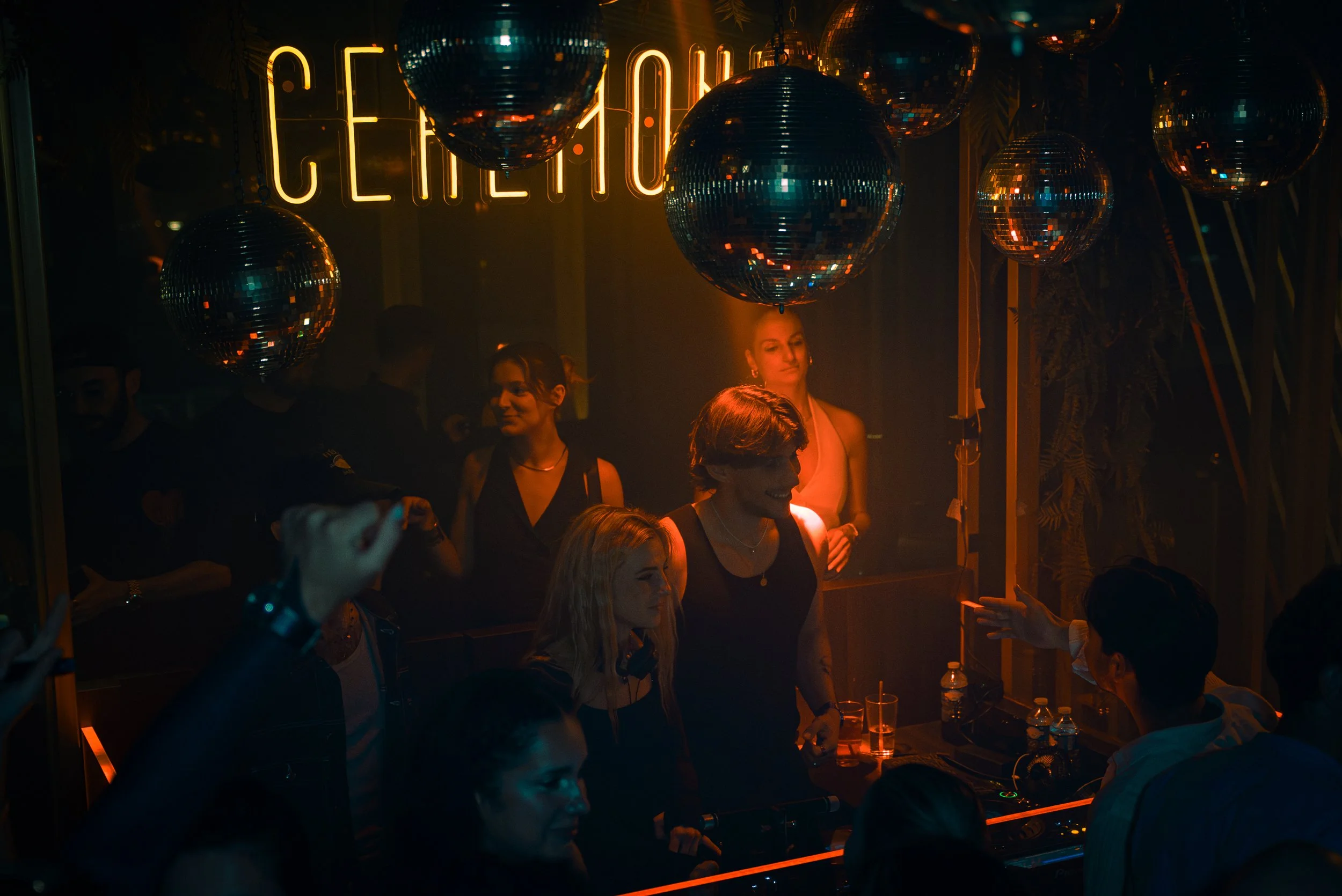 Une scène de soirée dans un club ou un bar, avec des personnes qui dansent et discutent, décorée avec des boules disco suspendues et un luminaire en néon "Centrik" en arrière-plan.