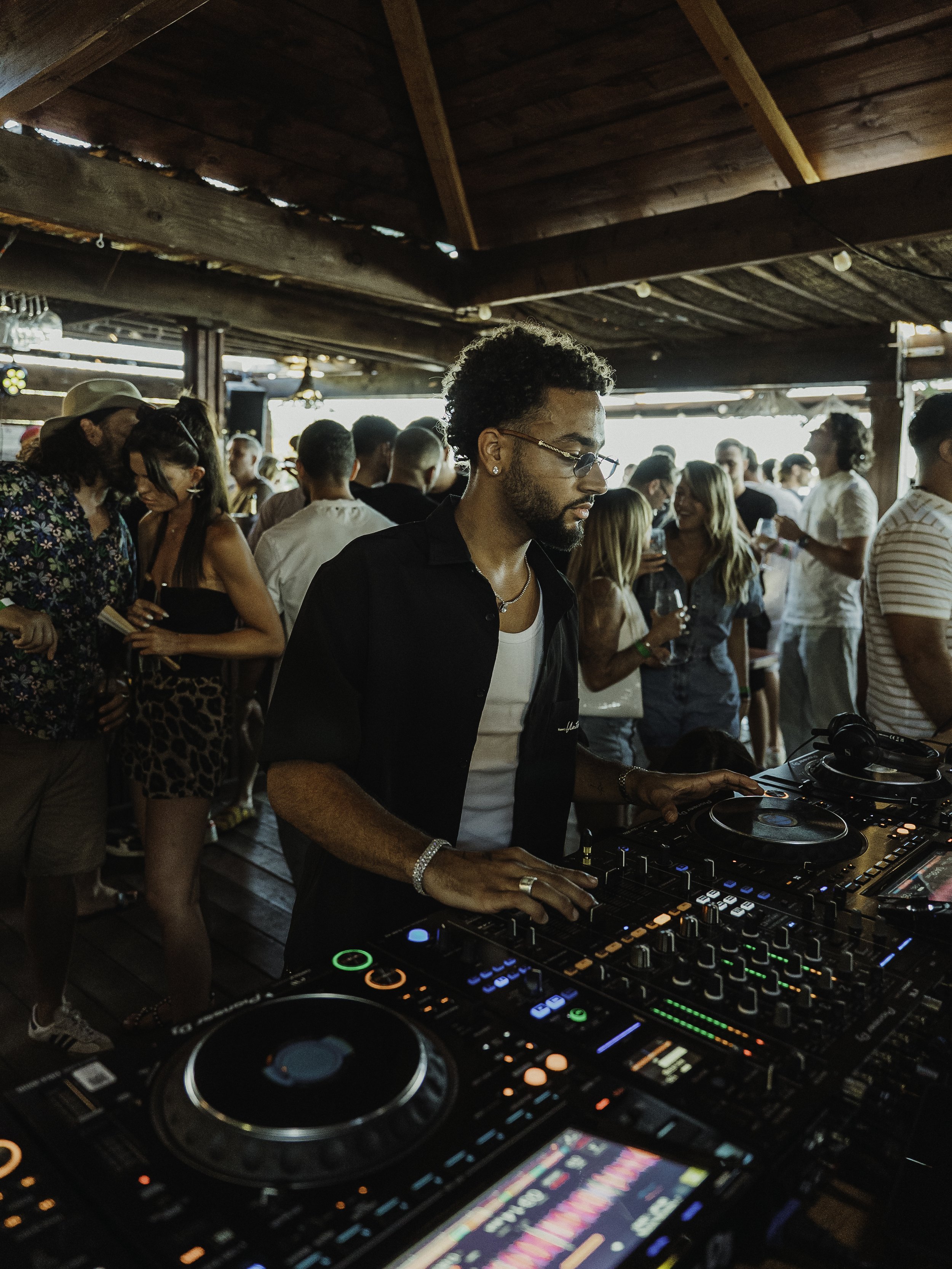 DJ mixant de la musique lors d'une fête ou d'un événement, entouré de plusieurs personnes qui dansent et socialisent dans une ambiance chaleureuse et festive.