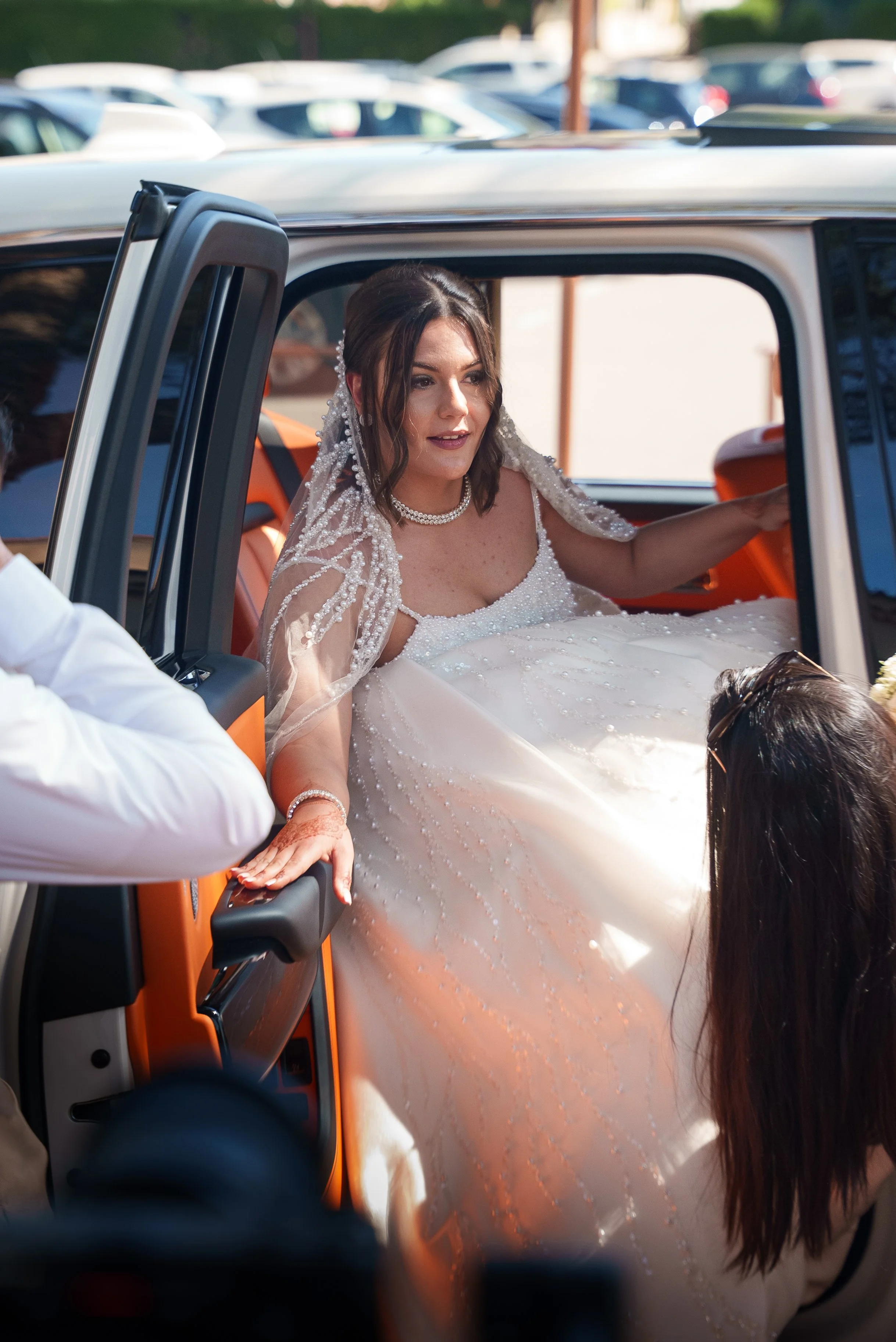 Une mariée en robe de mariage blanche sort d'un véhicule pendant un mariage, assistée par des personnes.