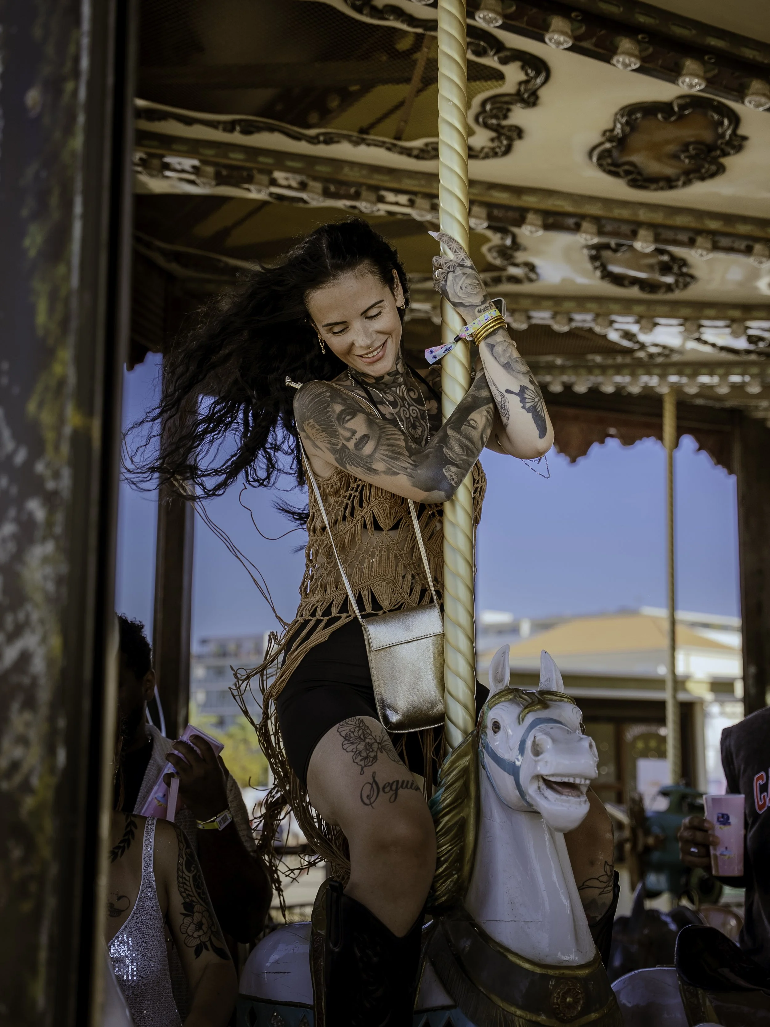 Une femme souriante avec des tatouages est assise sur un carrousel à cheval blanc, tenant une tige verticale, lors d'une fête en plein air sous un ciel bleu.