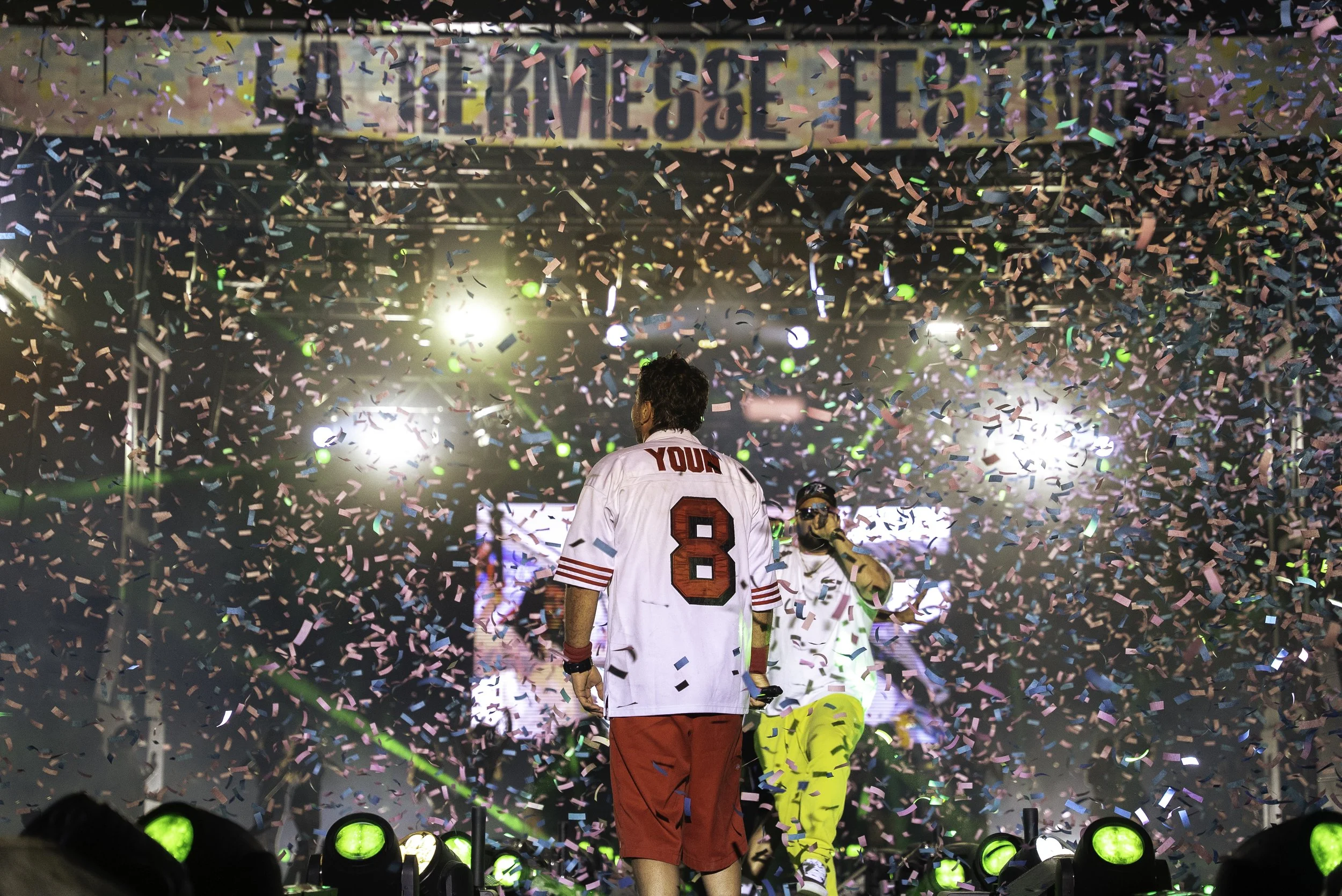 Scène de concert ou d'événement avec trois hommes sur scène, entourés de confettis colorés en suspension, avec des lumières vives et une grande bannière en arrière-plan.