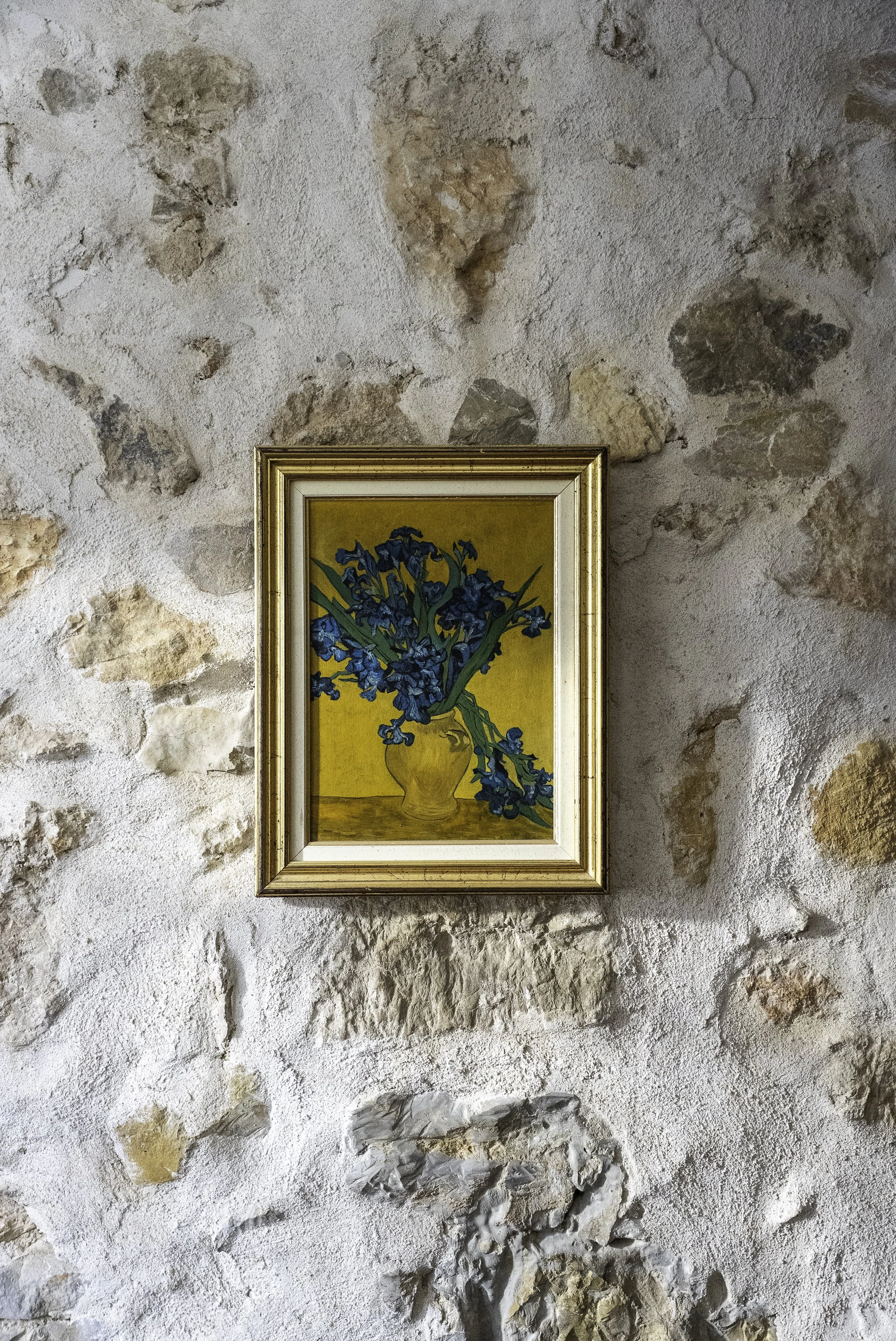Peinture représentant un vase avec des fleurs bleues, encadrée et suspendue à un mur en pierre.