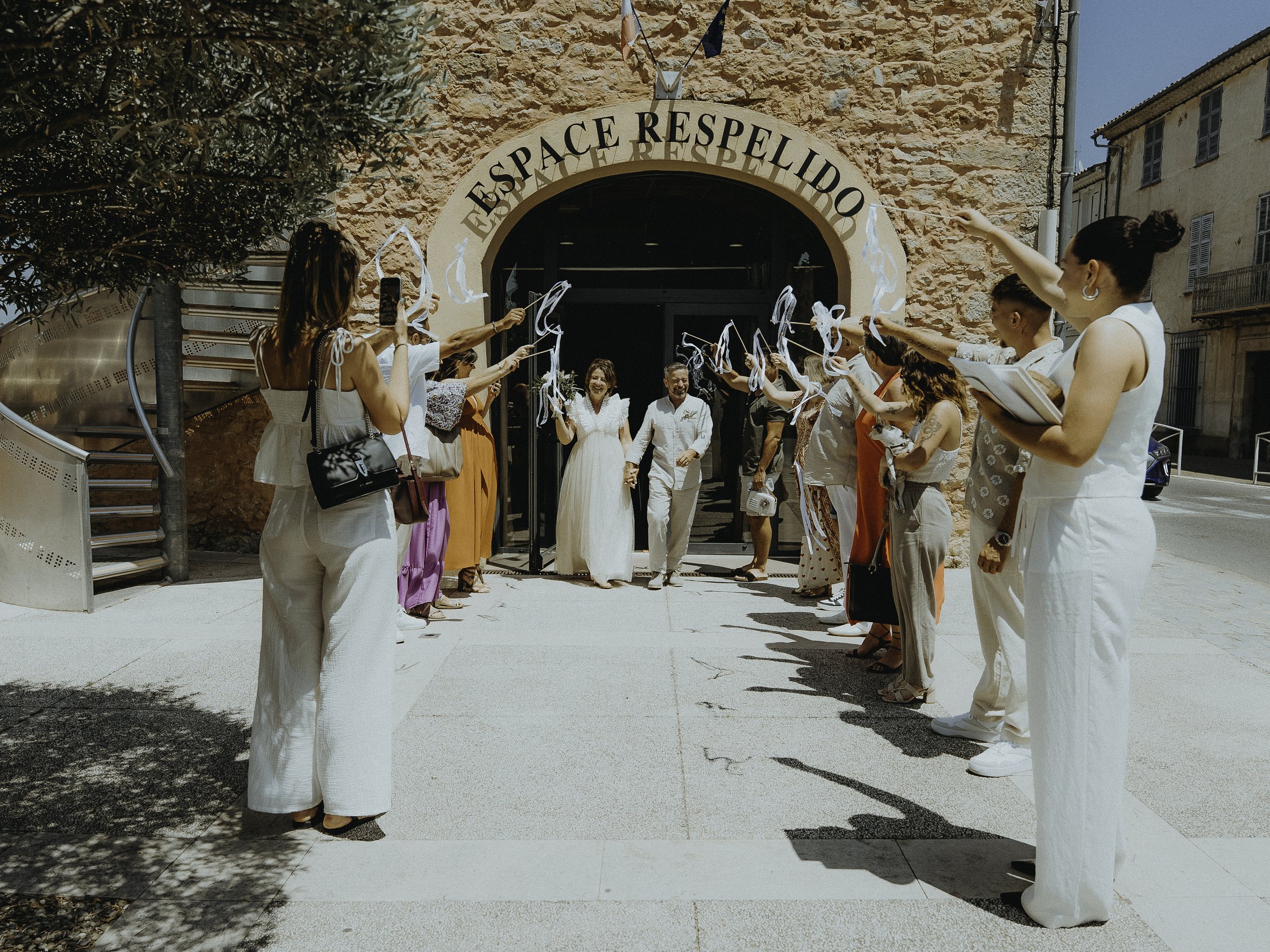 Un groupe de personnes célèbre le mariage d'une couple devant une entrée de bâtiment avec une arche. Les invités tiennent des rubans blancs lors d'une sortie de mariage en plein air, sous un ciel bleu. La scène est festive et ensoleillée.