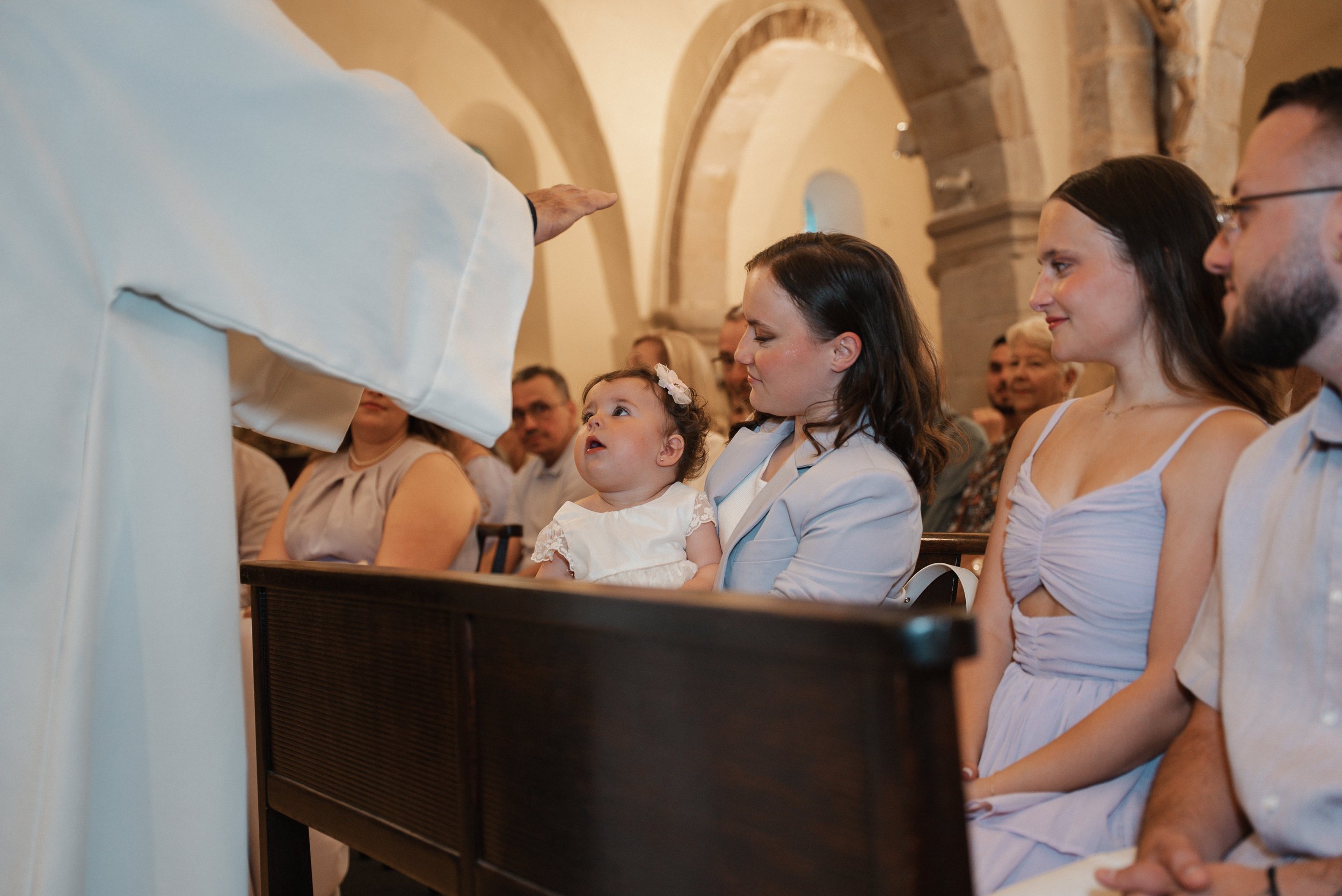 Une cérémonie religieuse dans une église, un prêtre bénit une famille assise dans les bancs, notamment une petite fille avec une femme vêtue de blanc, entourés d'autres adultes.