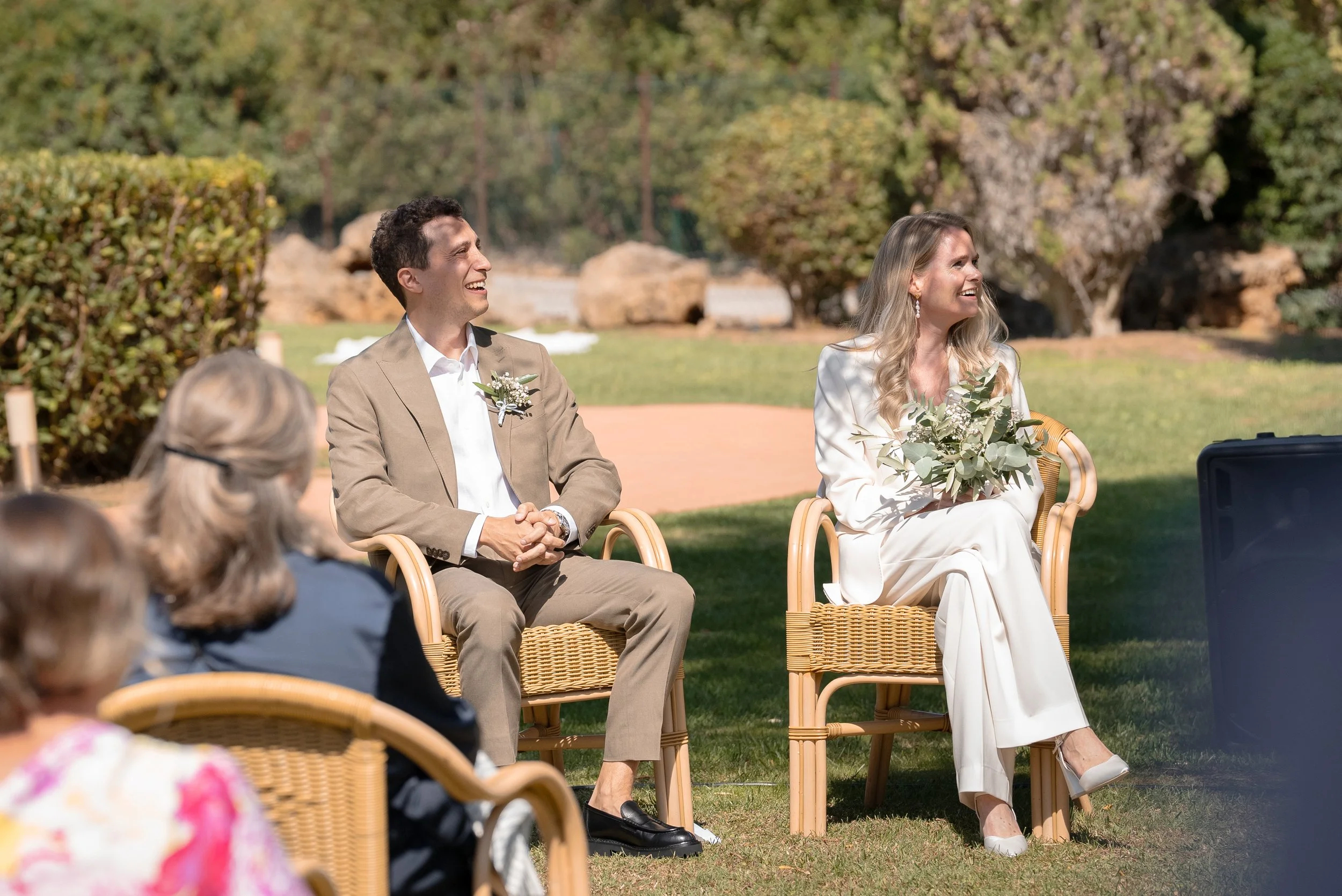 Un couple de mariés assis en plein air, la femme tient un bouquet de fleurs, tous deux sourient et rient lors d'une cérémonie de mariage, avec des invités en arrière-plan.