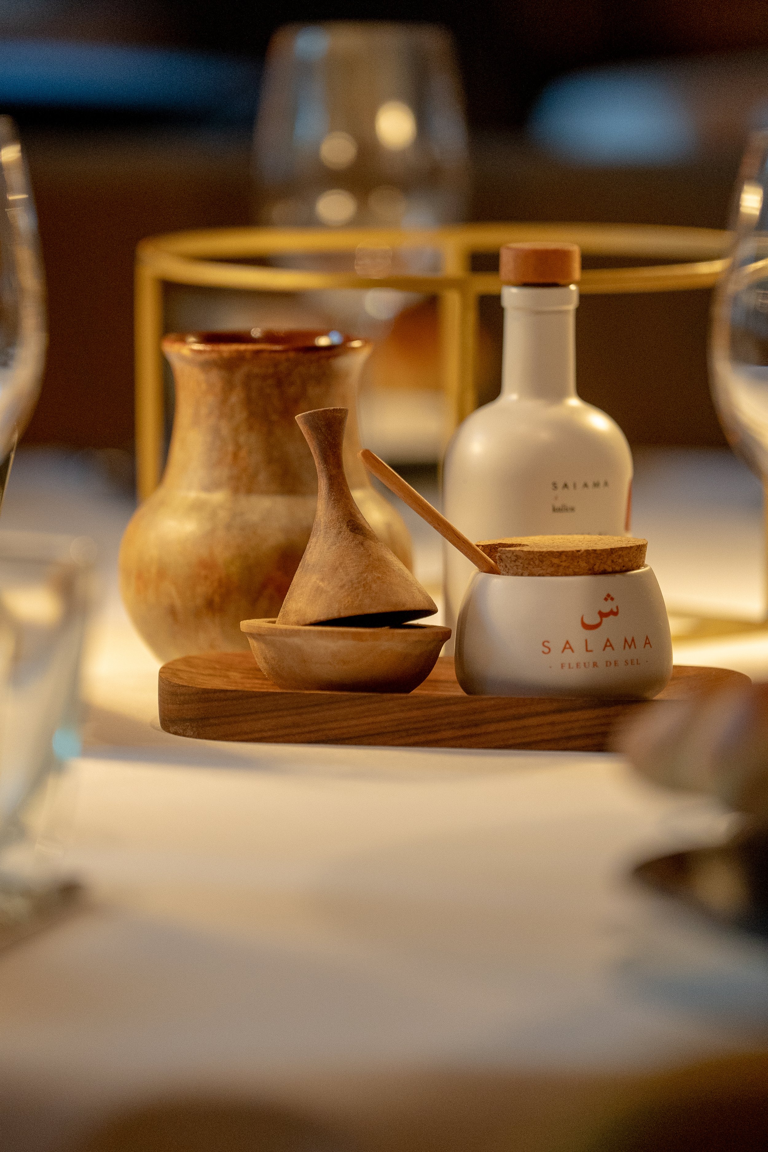 Objets décoratifs et épices sur une table, incluant un bocal blanc de sel de mer "Salama", un petit récipient en céramique, et un petit ustensile en bois, avec des verres sur la table en arrière-plan.