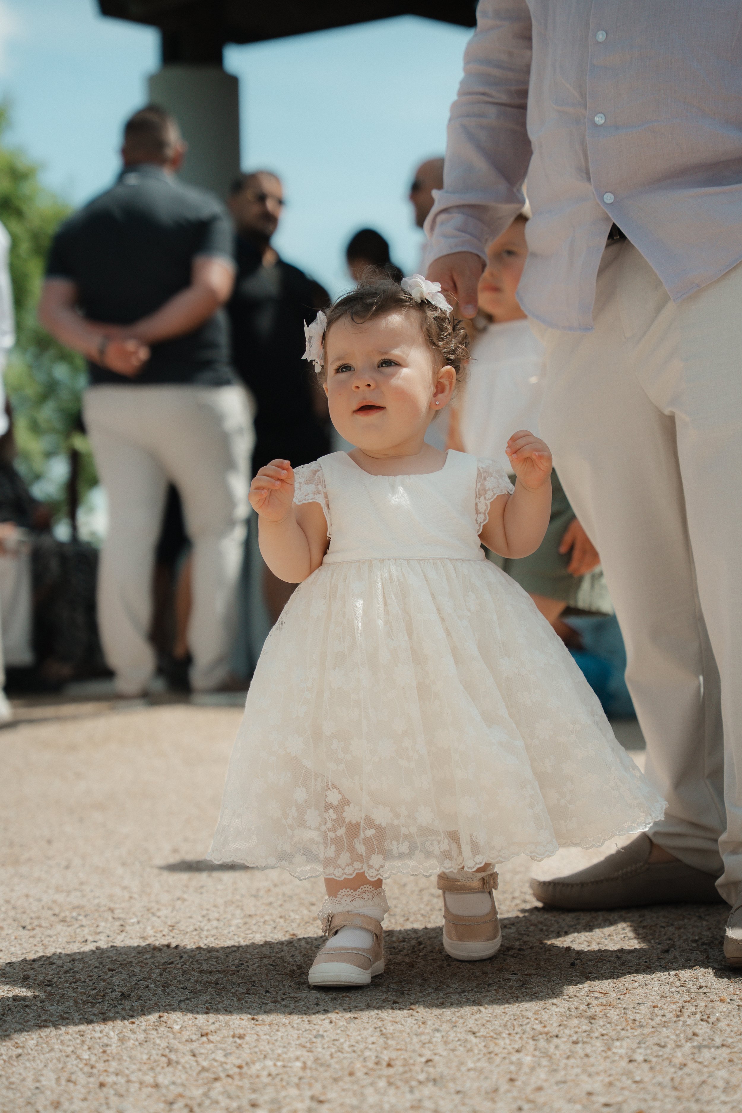 Jeune fille en robe blanche lors d'une cérémonie en extérieur, entourée d'adultes, sous un ciel clair et ensoleillé.